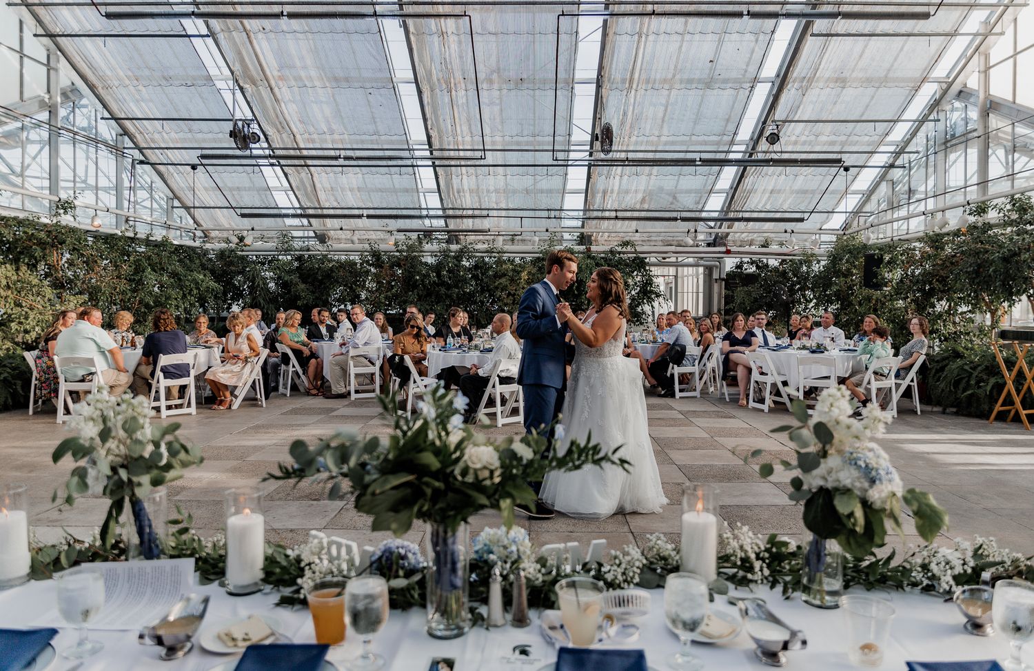 Wedding reception in a greenhouse venue with tables decorated with greenery and floral centerpieces.