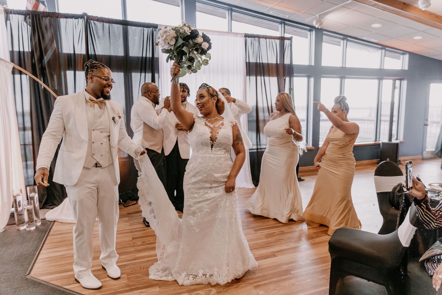 Wedding party celebrates joyfully on the dance floor in elegant white and tan attire.