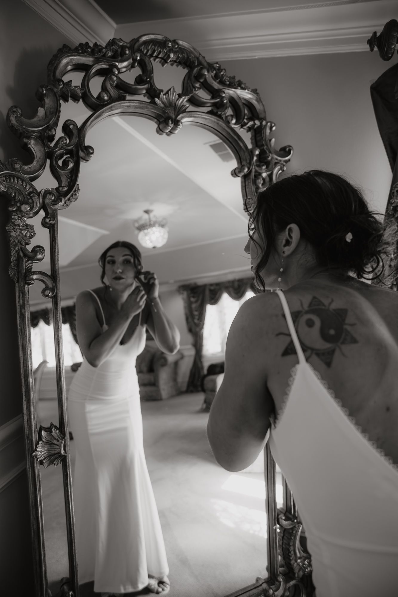 Bride in white dress looks in ornate vintage mirror while applying makeup in black and white photo.