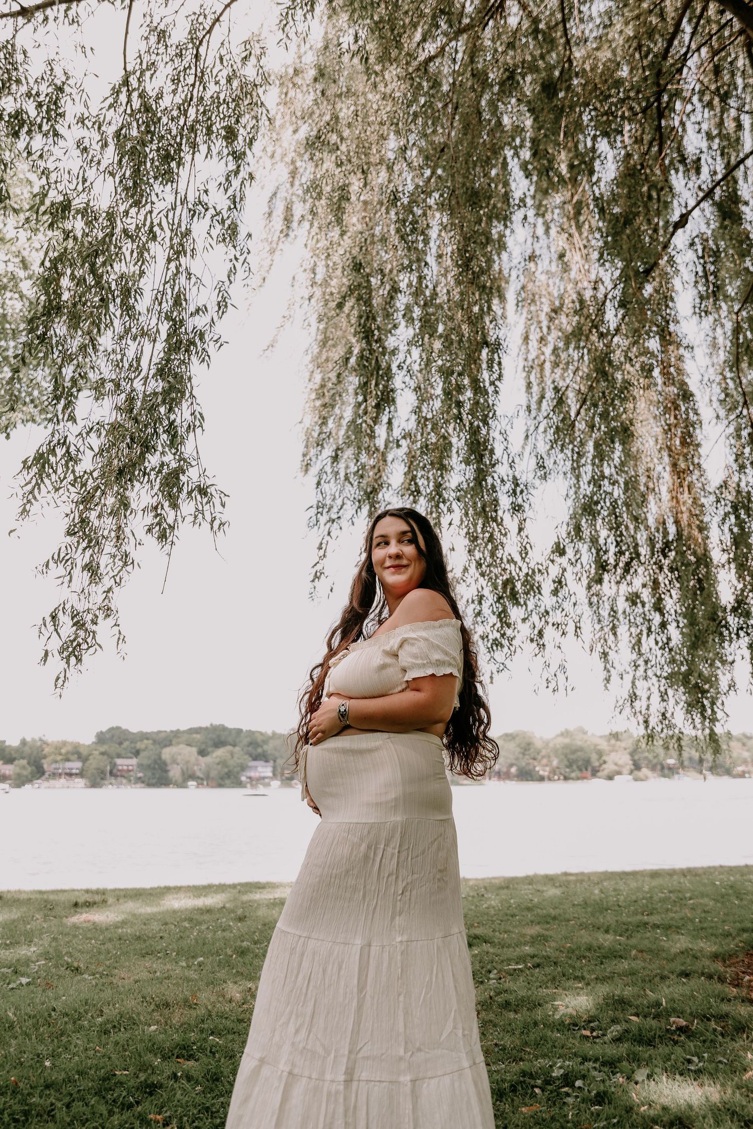 Woman in white off-shoulder maxi dress smiling under weeping willow tree by lakeside on a cloudy day.