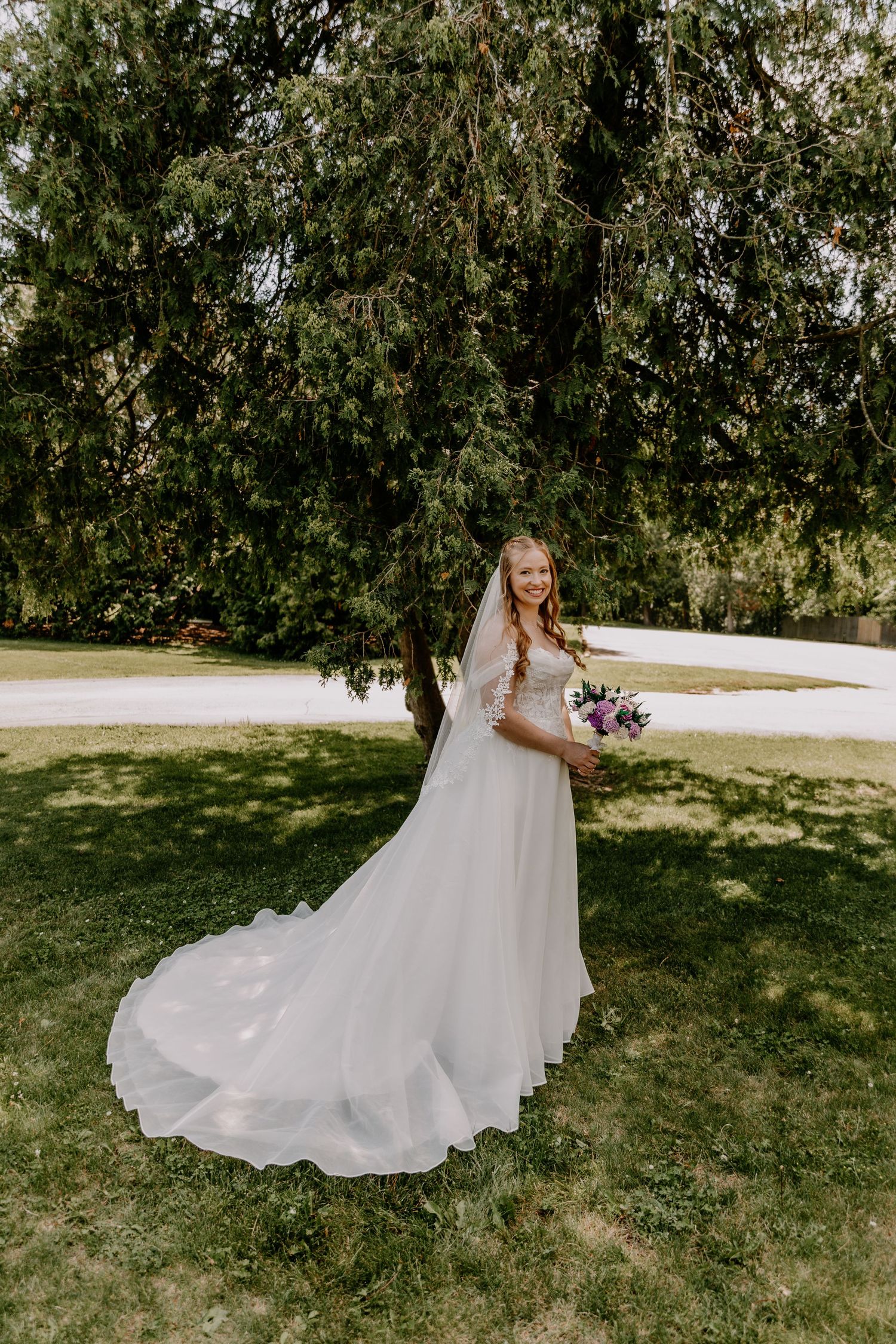 Smiling bride in a long white gown and veil holds a purple bouquet outdoors under a large green tree.