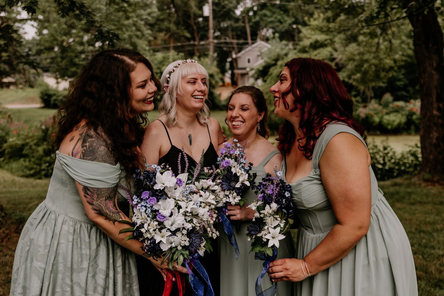 Four bridesmaids in sage green dresses laughing together outdoors, holding purple and white floral bouquets at a wedding.