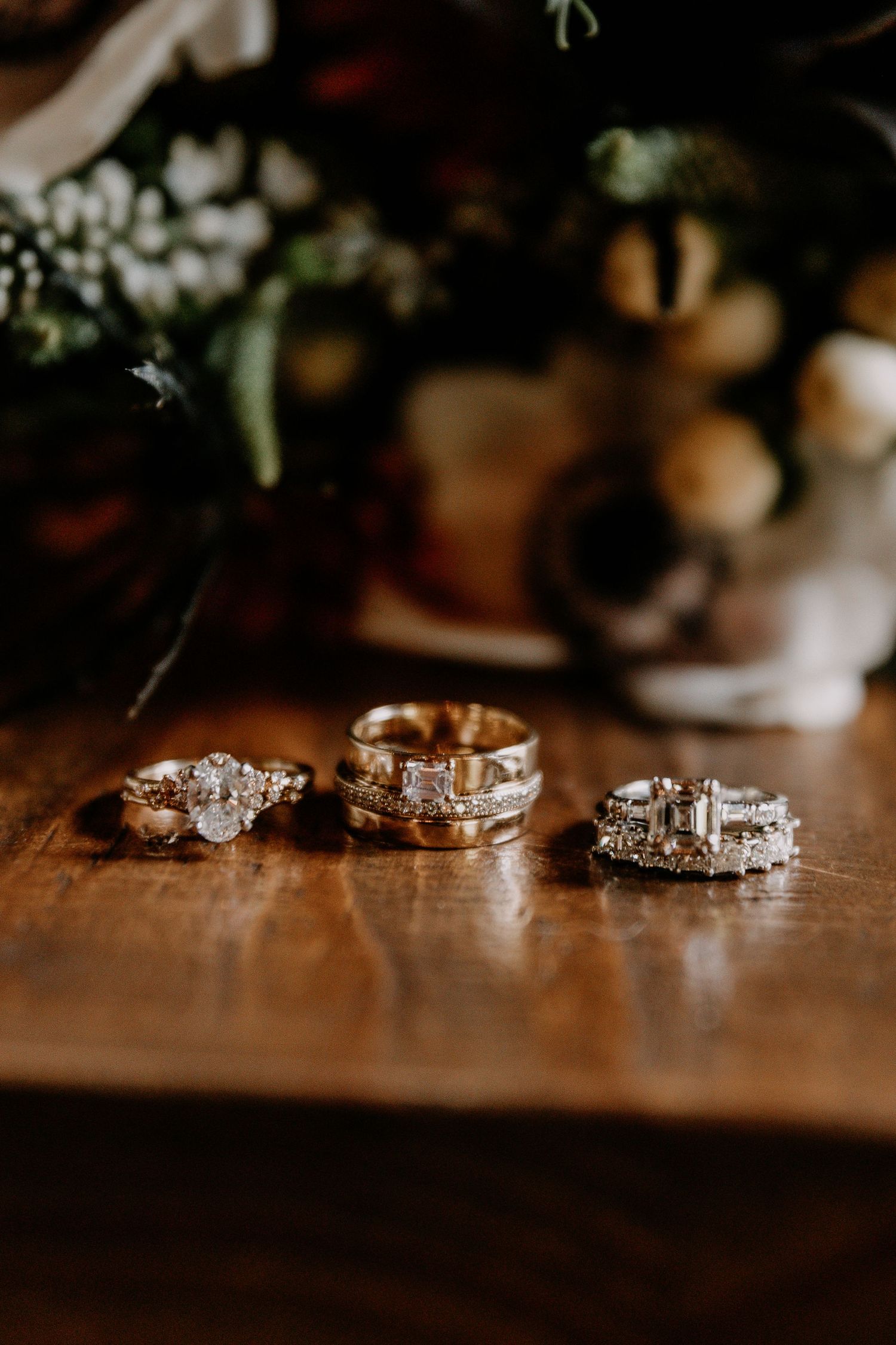 Three elegant wedding rings with diamond details arranged on a wooden surface with flowers in the background.