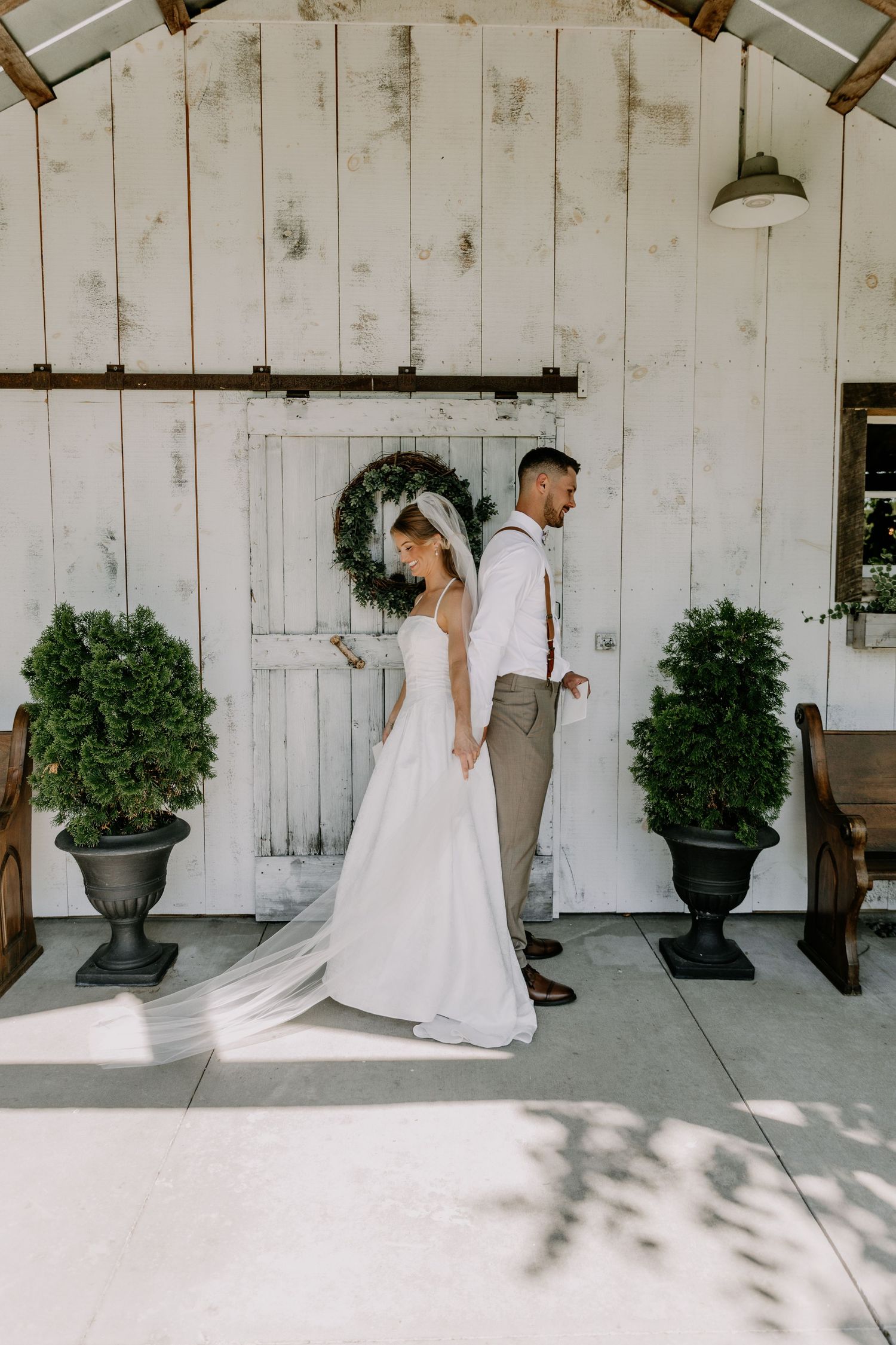 Bride and groom stand back-to-back before a rustic white barn door adorned with a greenery wreath on their wedding day.