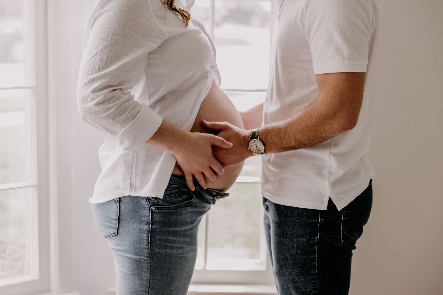 Pregnant couple standing by window, man placing hands on woman's baby bump, both wearing white shirts and jeans.