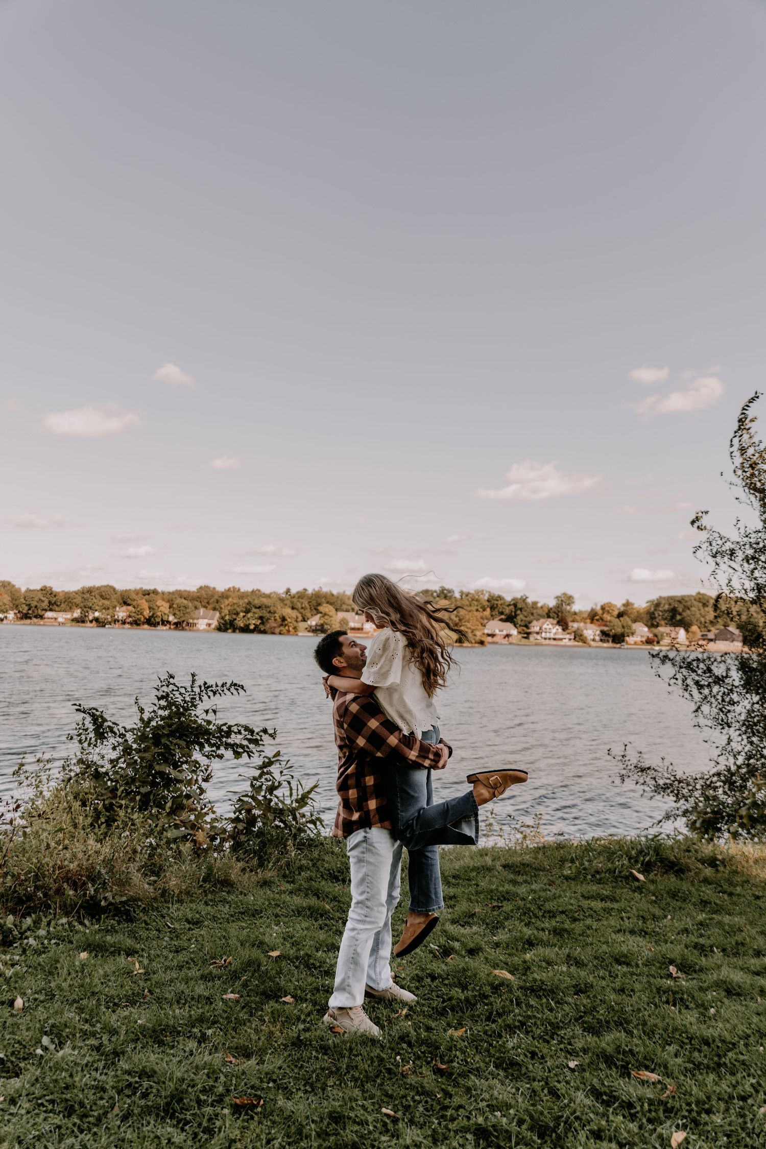 Couple embracing by a lakeside, man lifting woman in a joyful moment, surrounded by lush greenery and calm water.
