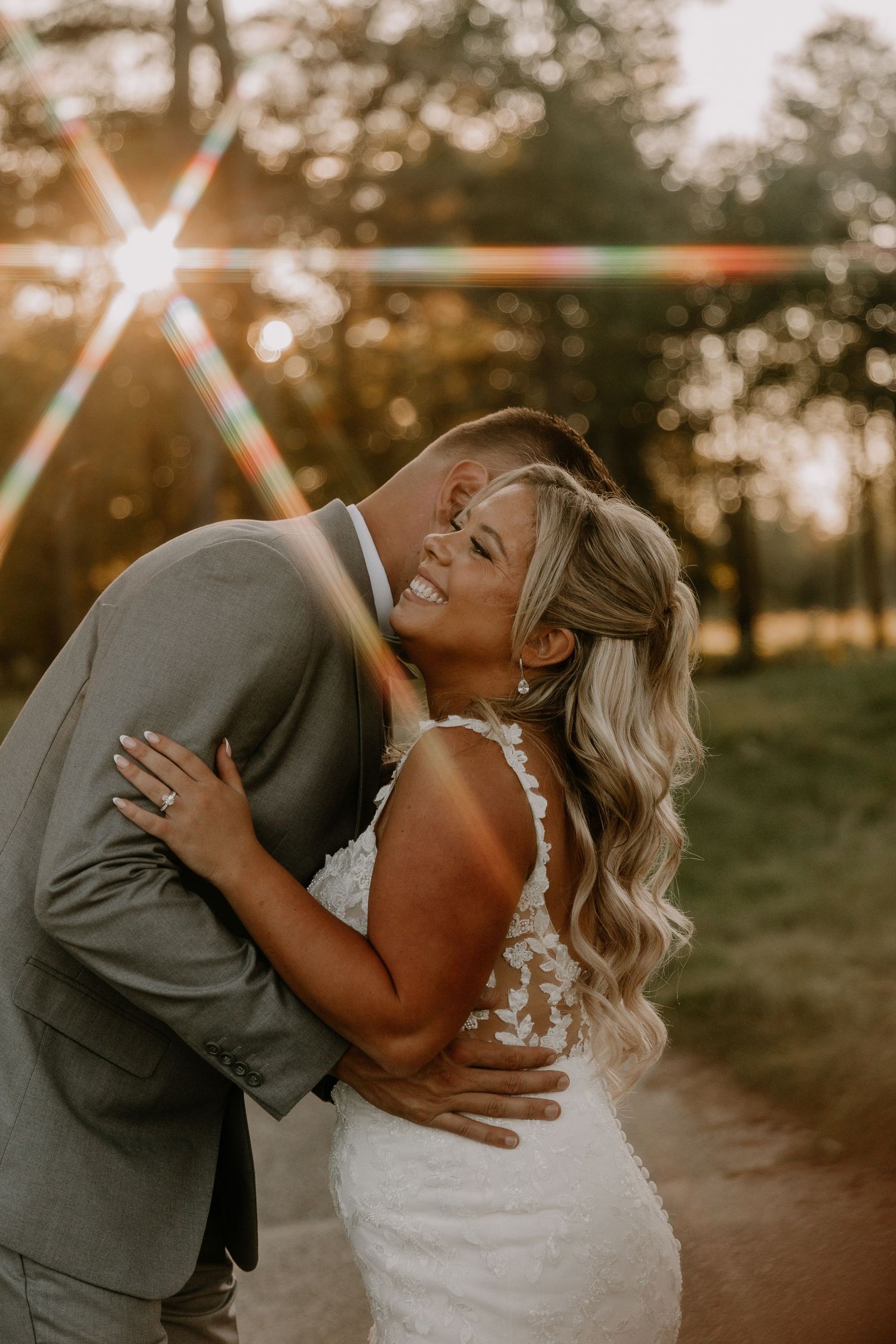 A bride in lace gown embraces her groom in gray suit during golden hour sunset at outdoor wedding.