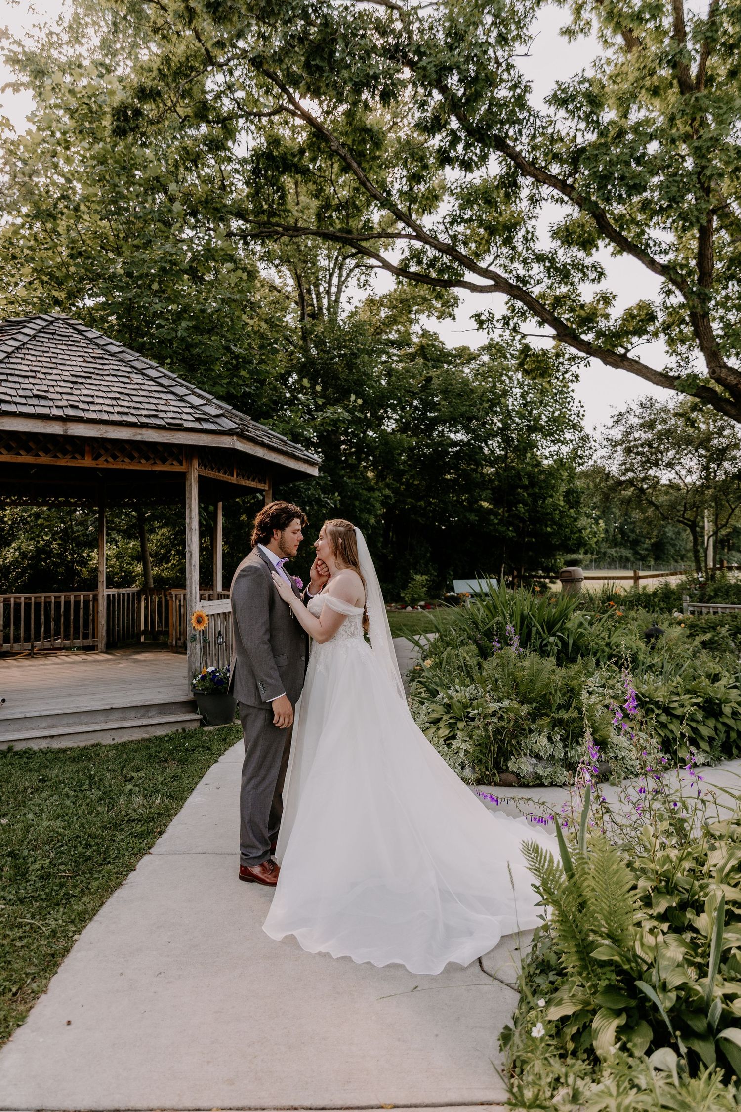 A bride and groom share a romantic moment on a garden path near a gazebo, surrounded by lush greenery.