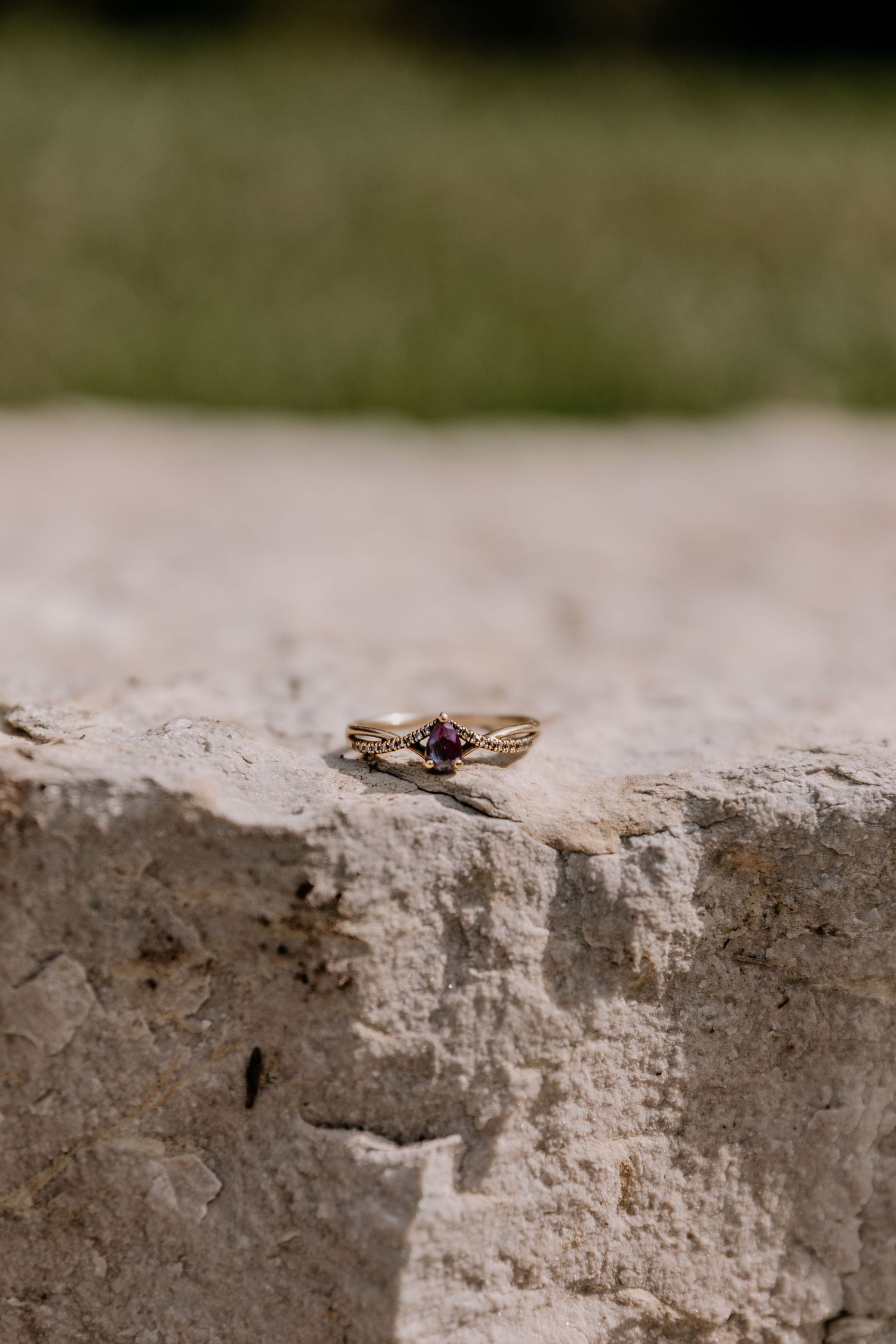 Gold engagement ring with dark red gemstone displayed on rustic stone surface with blurred green background.