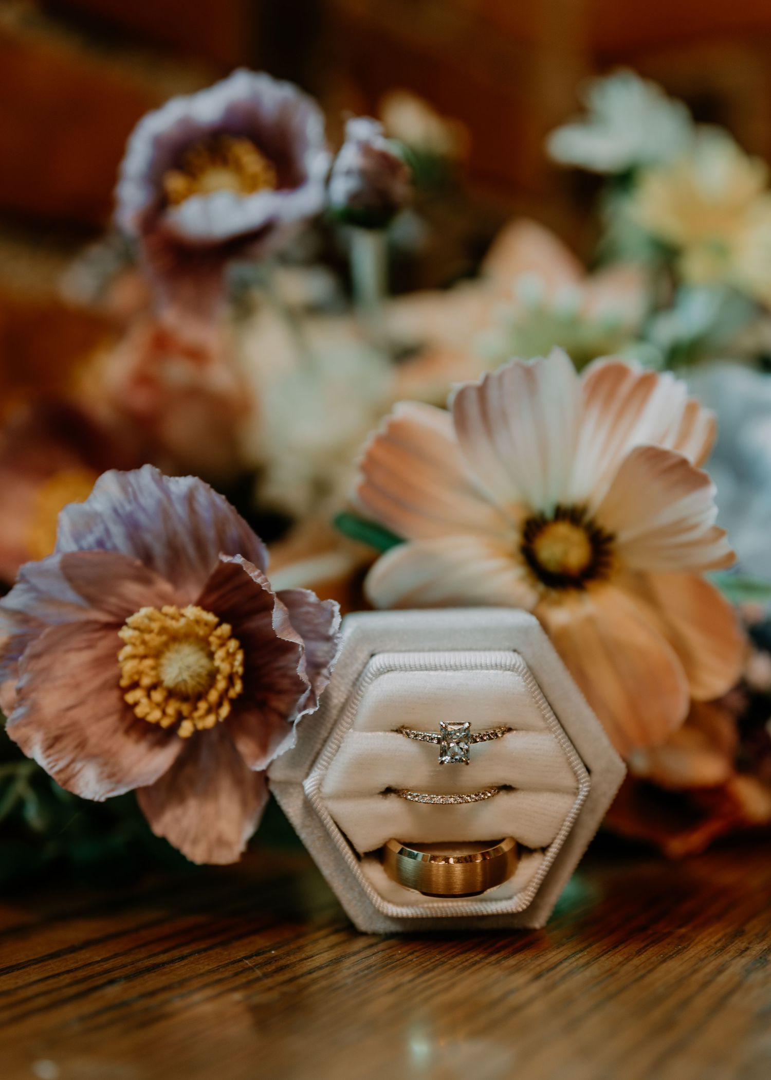 Wedding rings displayed in a hexagonal ring box surrounded by soft pink and peach cosmos flowers on a wooden surface.