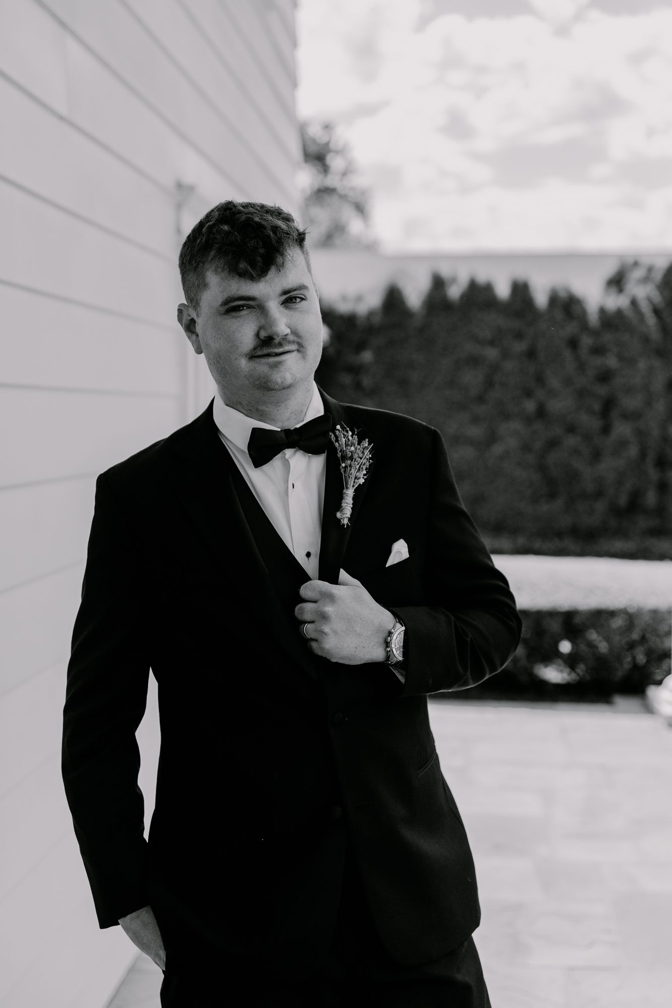 A black and white portrait of a groom in formal attire standing beside a white wall.