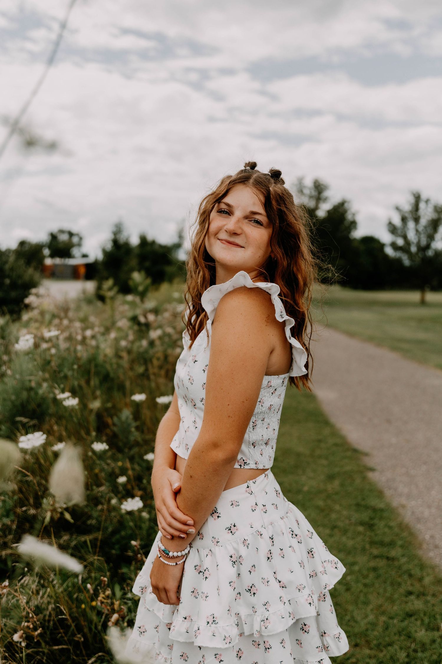 Woman in white floral dress poses on a path surrounded by wildflowers on a summer day.