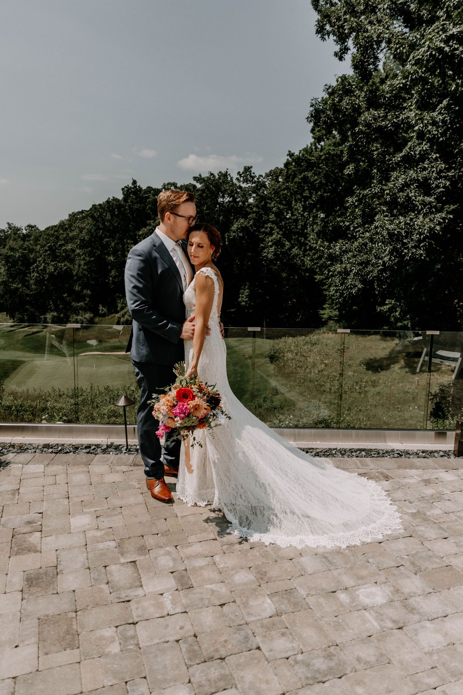 Wedding couple embracing on stone patio with bride holding colorful bouquet and wearing flowing lace dress.