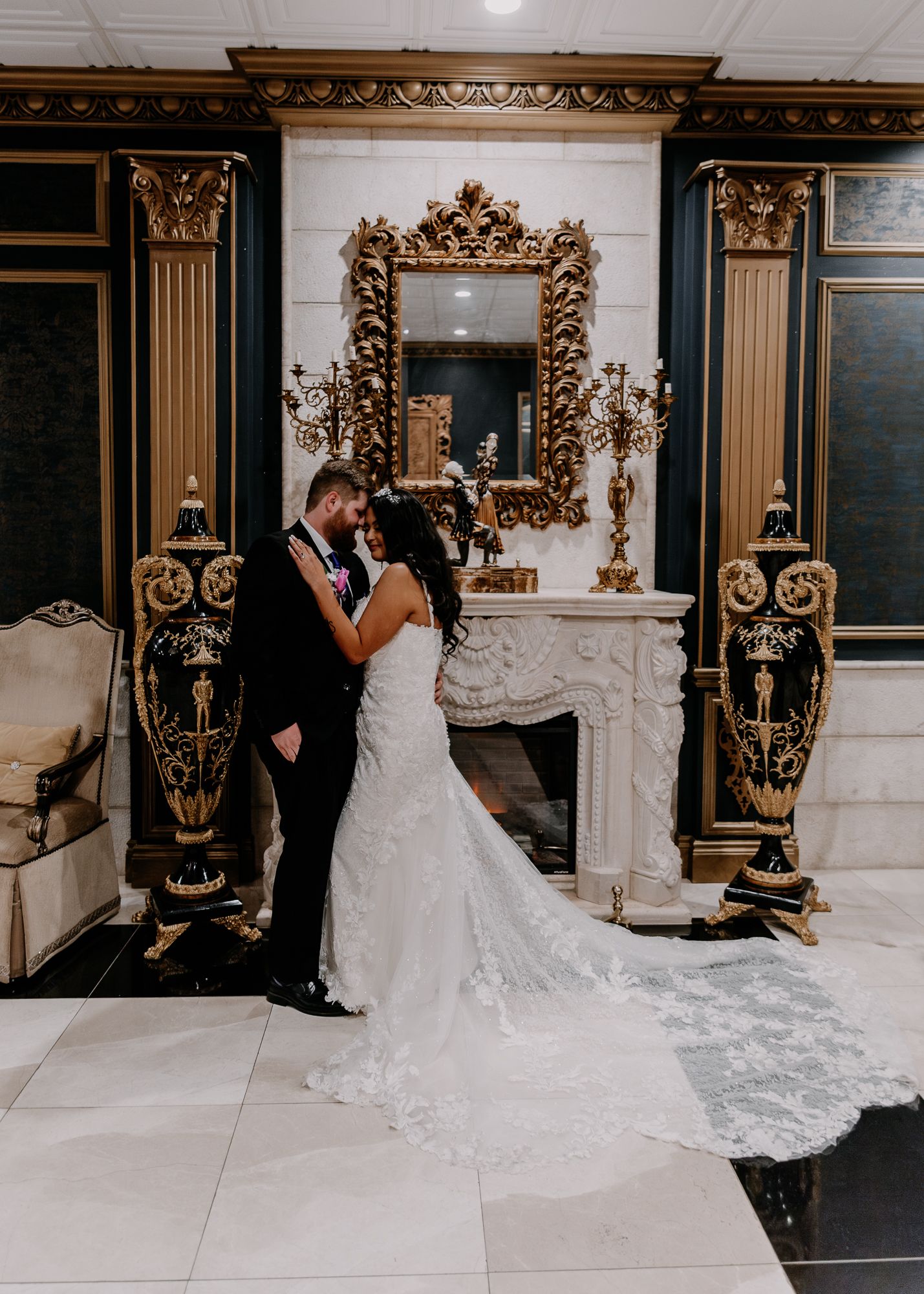 Elegant wedding portrait by ornate gold mirror and fireplace in classic interior setting.