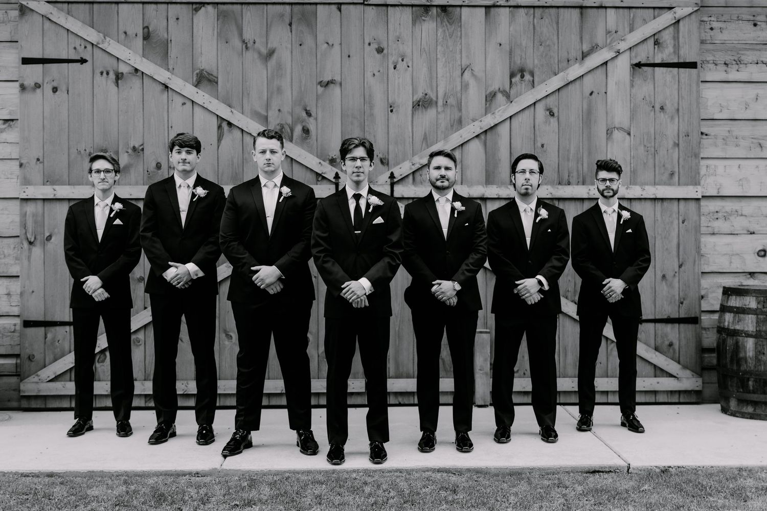 Groomsmen in black suits line up against barn door background for formal wedding photo.