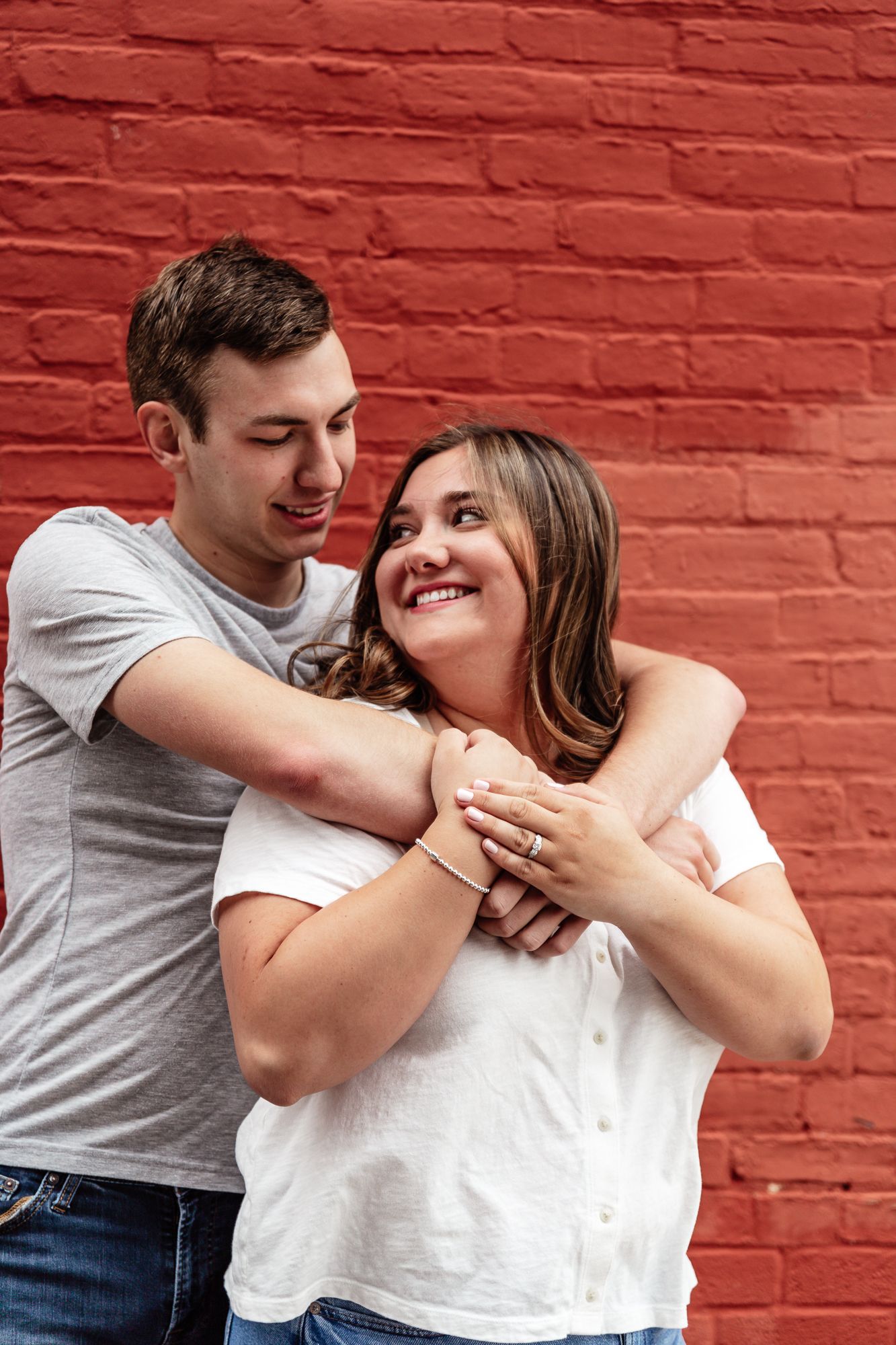 Couple laughs while embracing against vibrant red brick wall during photo session.