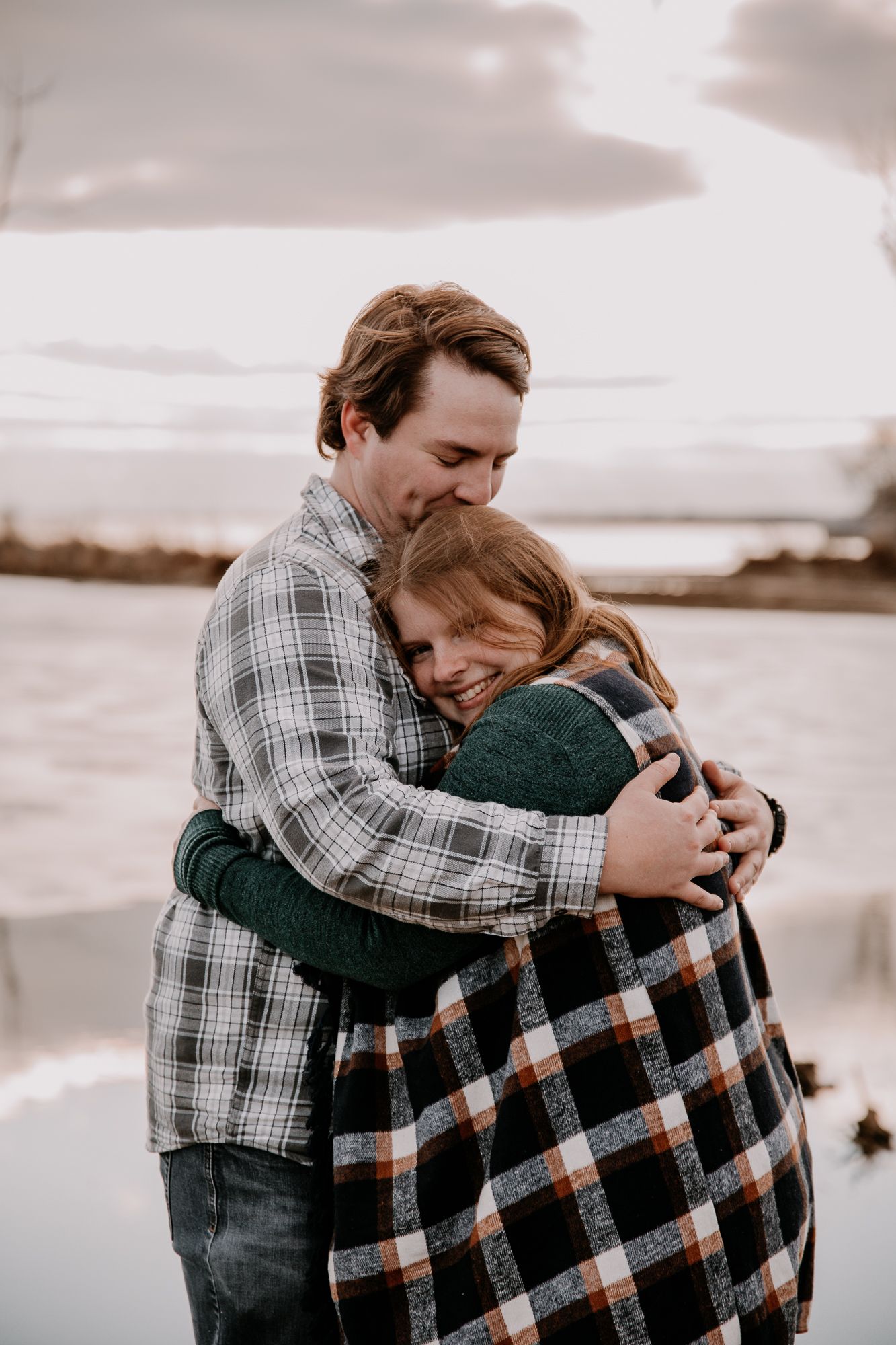 Two people share an intimate embrace while wrapped in a plaid blanket at sunset.