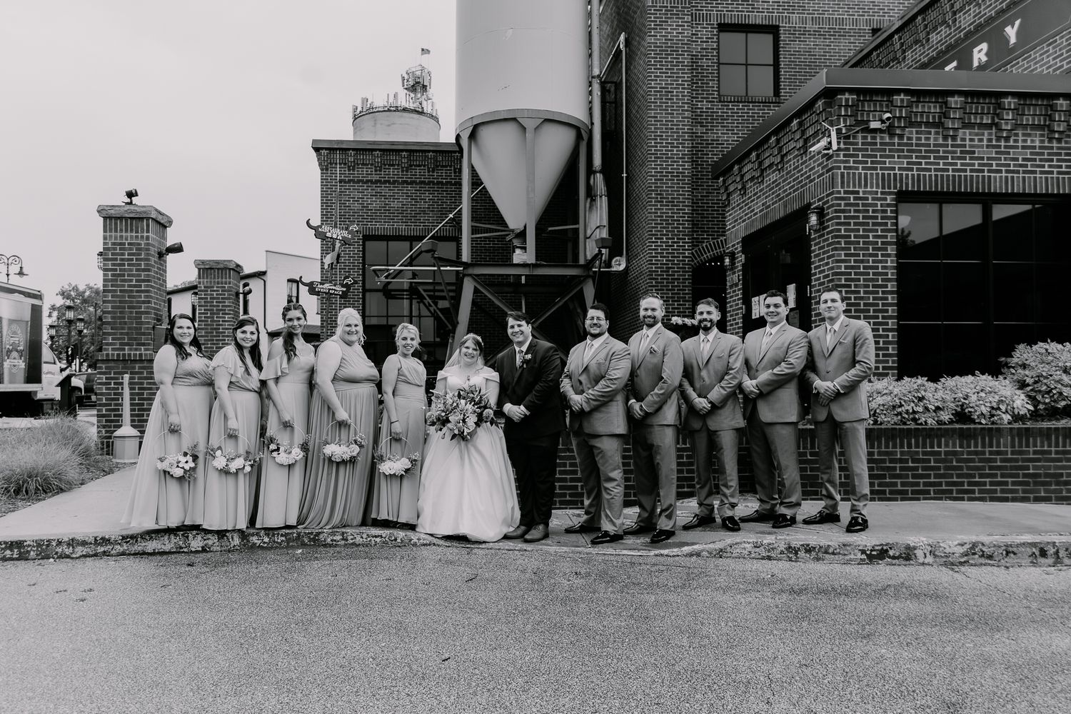 Black and white panorama shows wedding party lined up outside brick building.