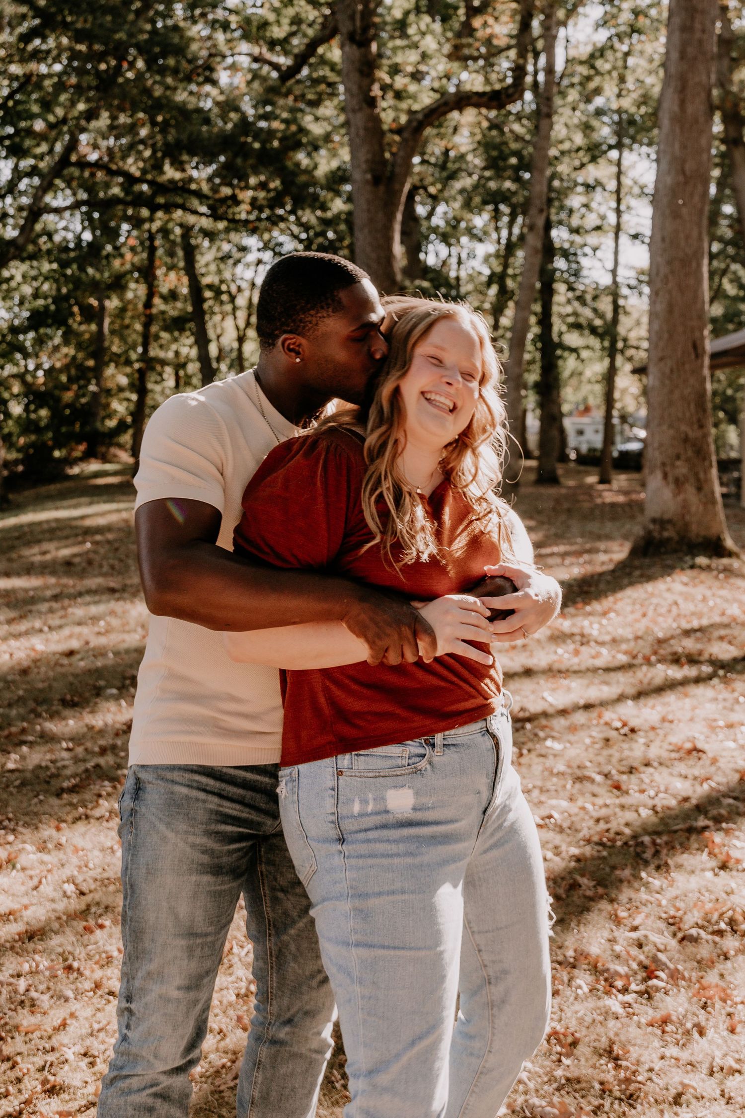 A couple embraces and shares affectionate moments in a sunlit wooded area on a warm autumn day.