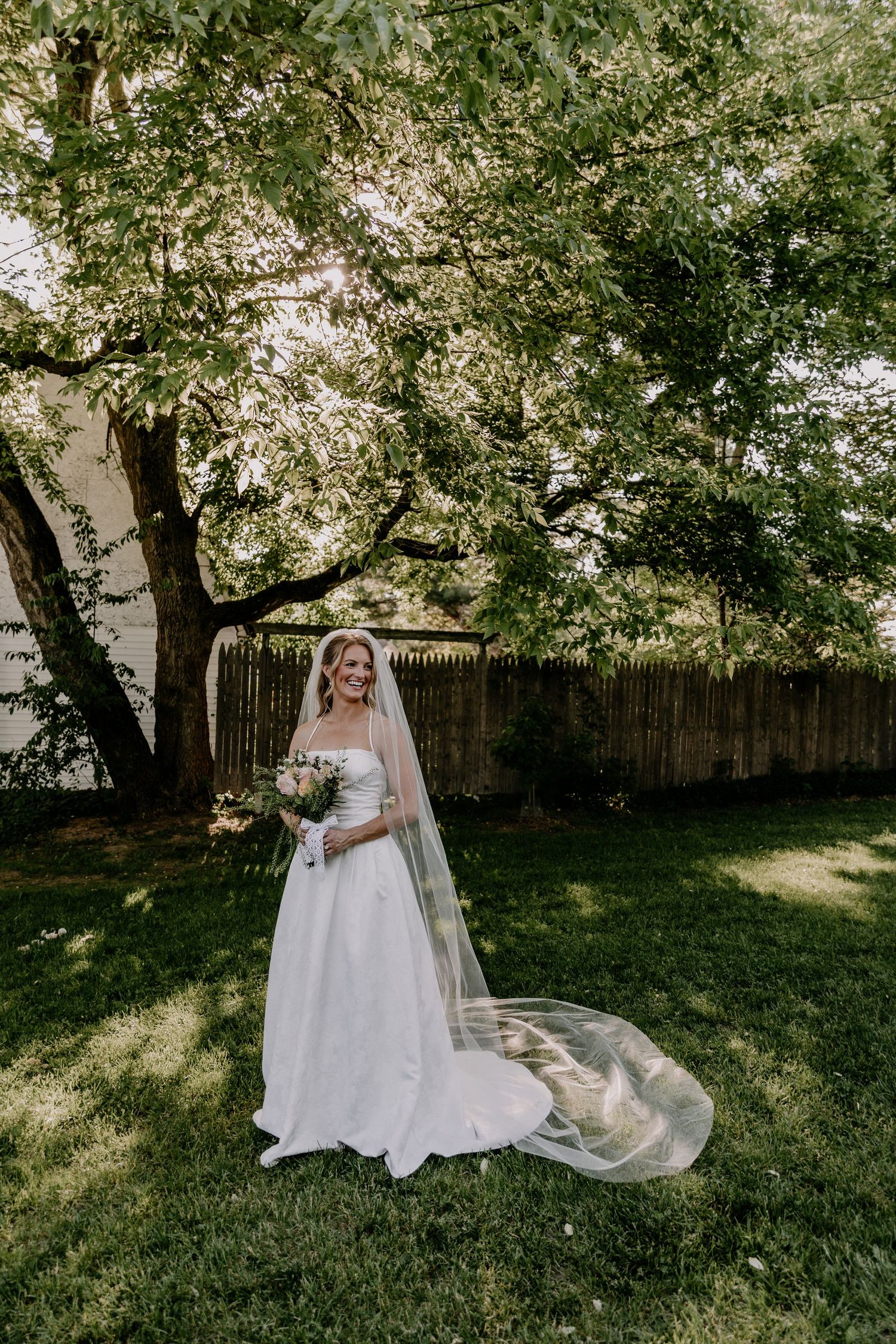 Smiling bride in a white ball gown and long veil holds a bouquet while standing on green grass beneath a large tree.
