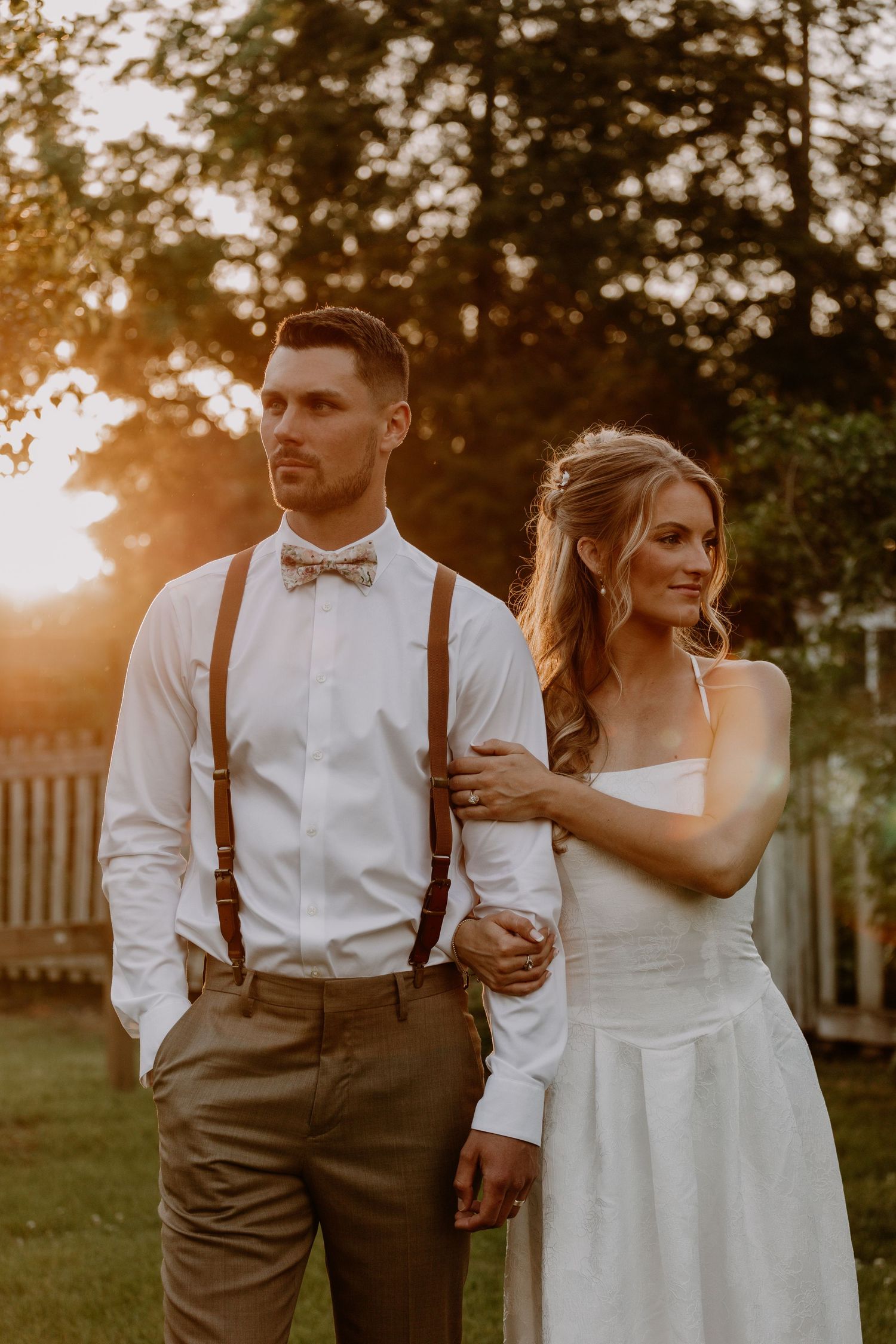 Bride and groom pose at golden hour sunset, groom in white shirt with suspenders and bow tie, bride in white dress.