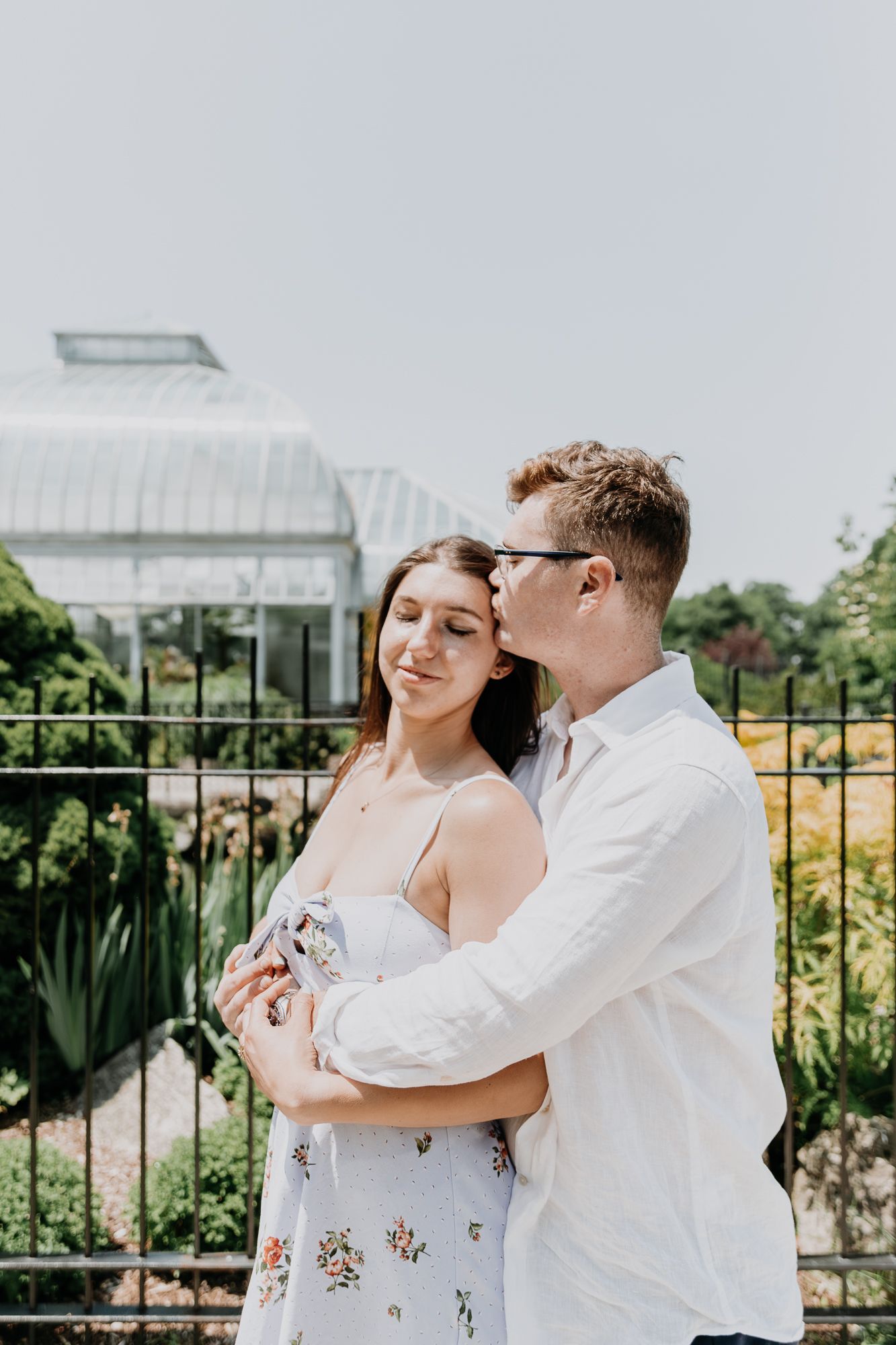 Two people share an intimate moment while standing near a greenhouse garden with flowering plants.