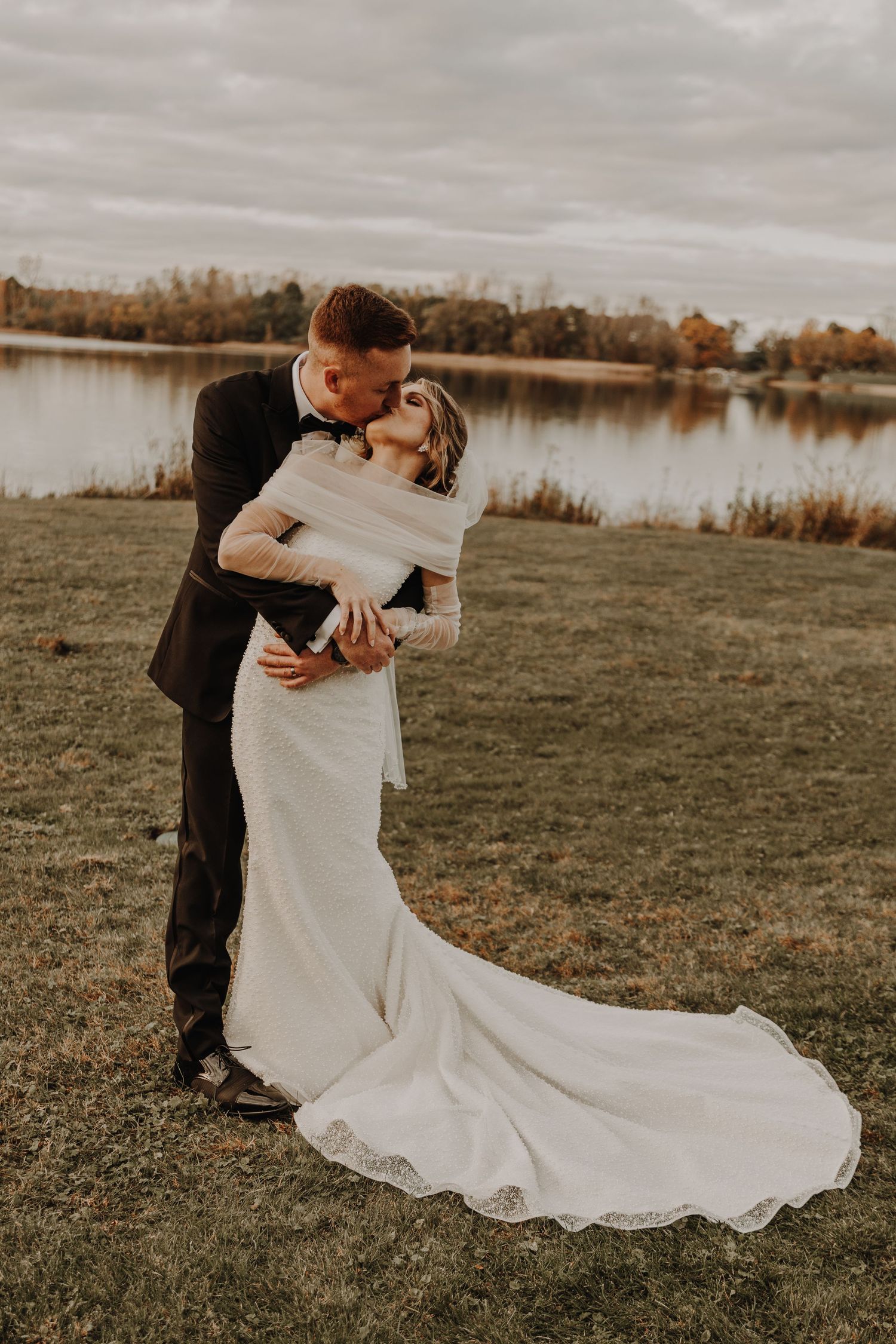 Wedding couple embraces in an elegant pose by a lake with the bride's dress trailing beautifully behind.
