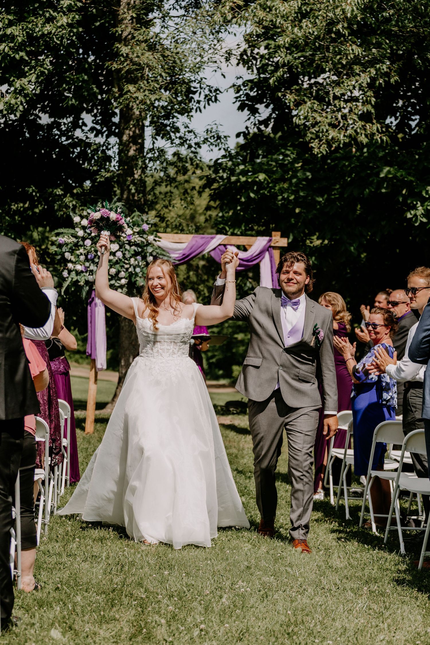 Joyful newlyweds raise hands walking down outdoor aisle, bride in white gown holding purple bouquet, groom in gray suit.