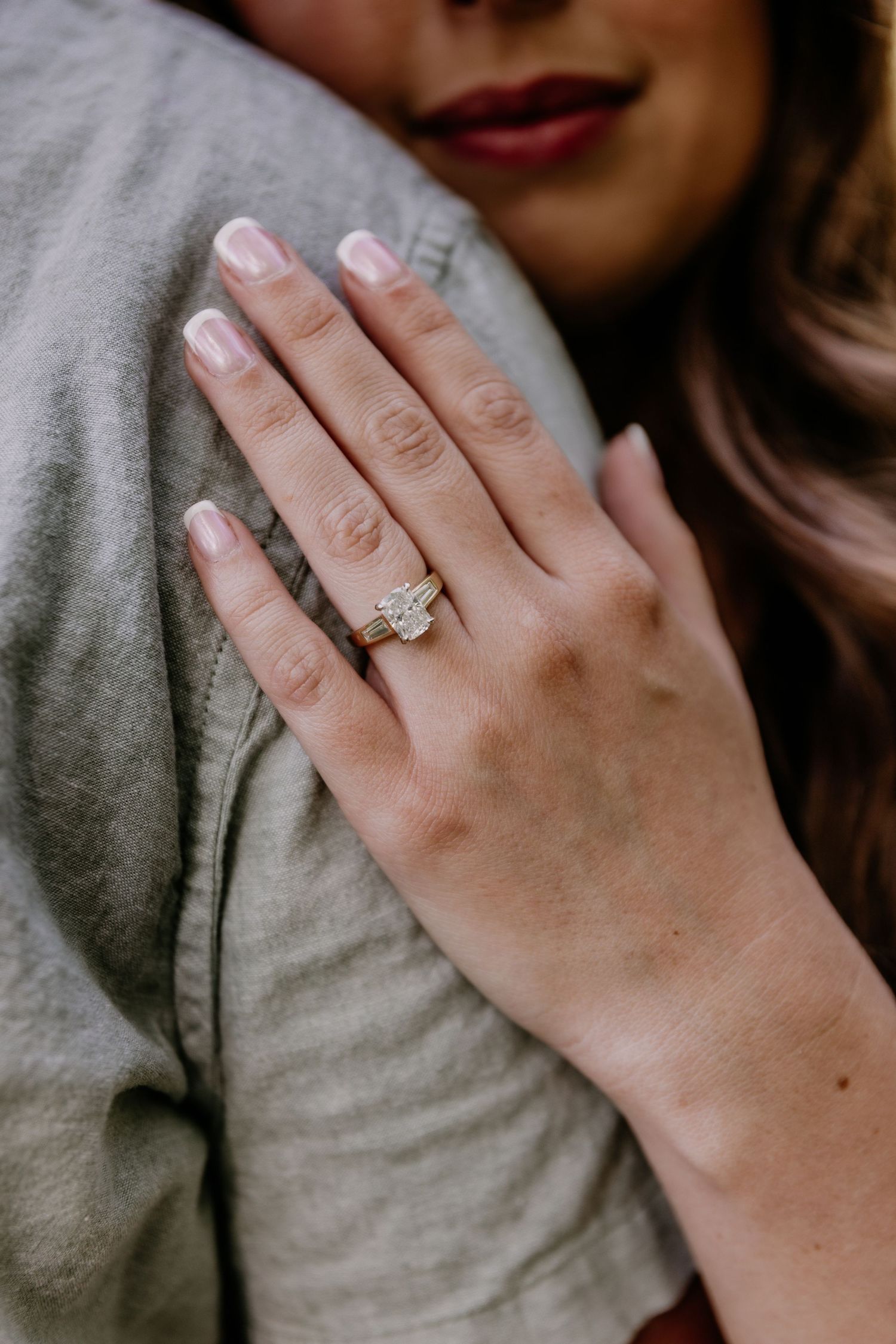 Woman displaying a stunning emerald-cut diamond engagement ring on gold band while embracing her partner.