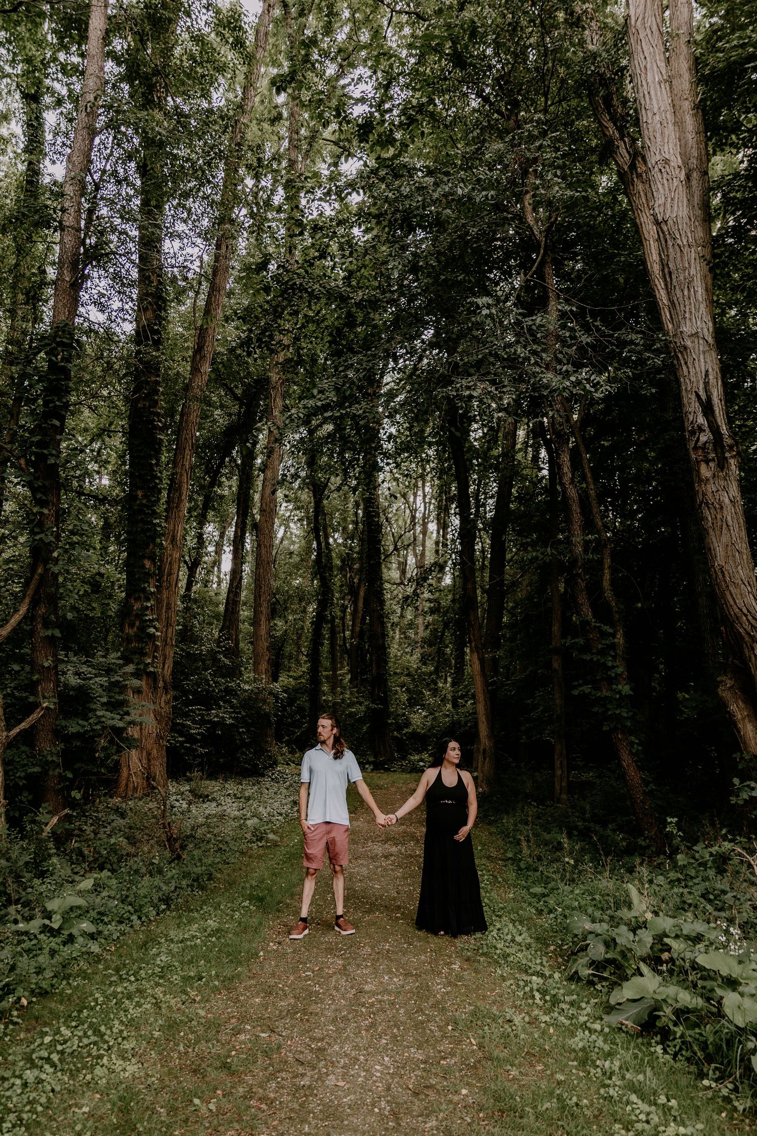 Couple holding hands on a forest path surrounded by tall green trees, man in pink shorts and woman in black dress.