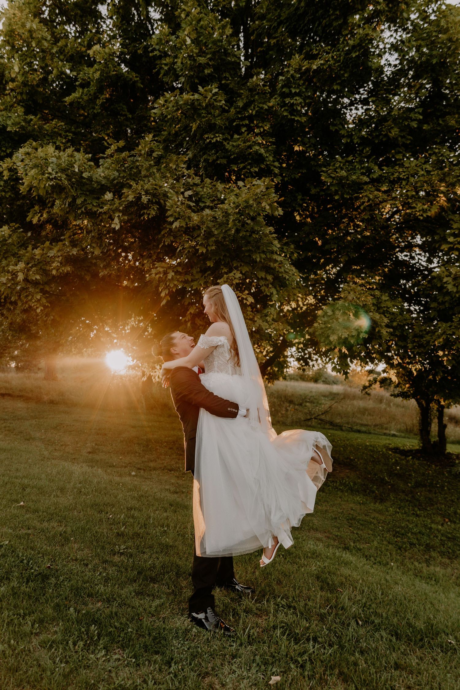 Groom lifts bride in white gown at golden sunset in lush green garden during romantic outdoor wedding celebration.