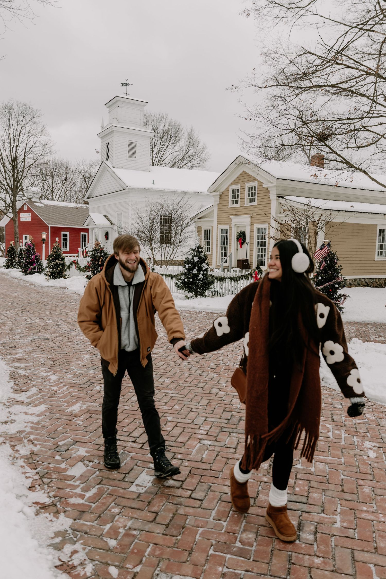 A winter photoshoot on a snowy brick path with colonial-style buildings and evergreens in the background.