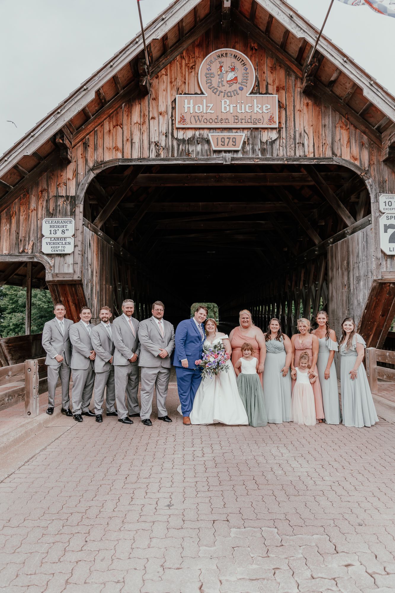 Wedding party poses in front of rustic covered bridge with bride and groom in center.
