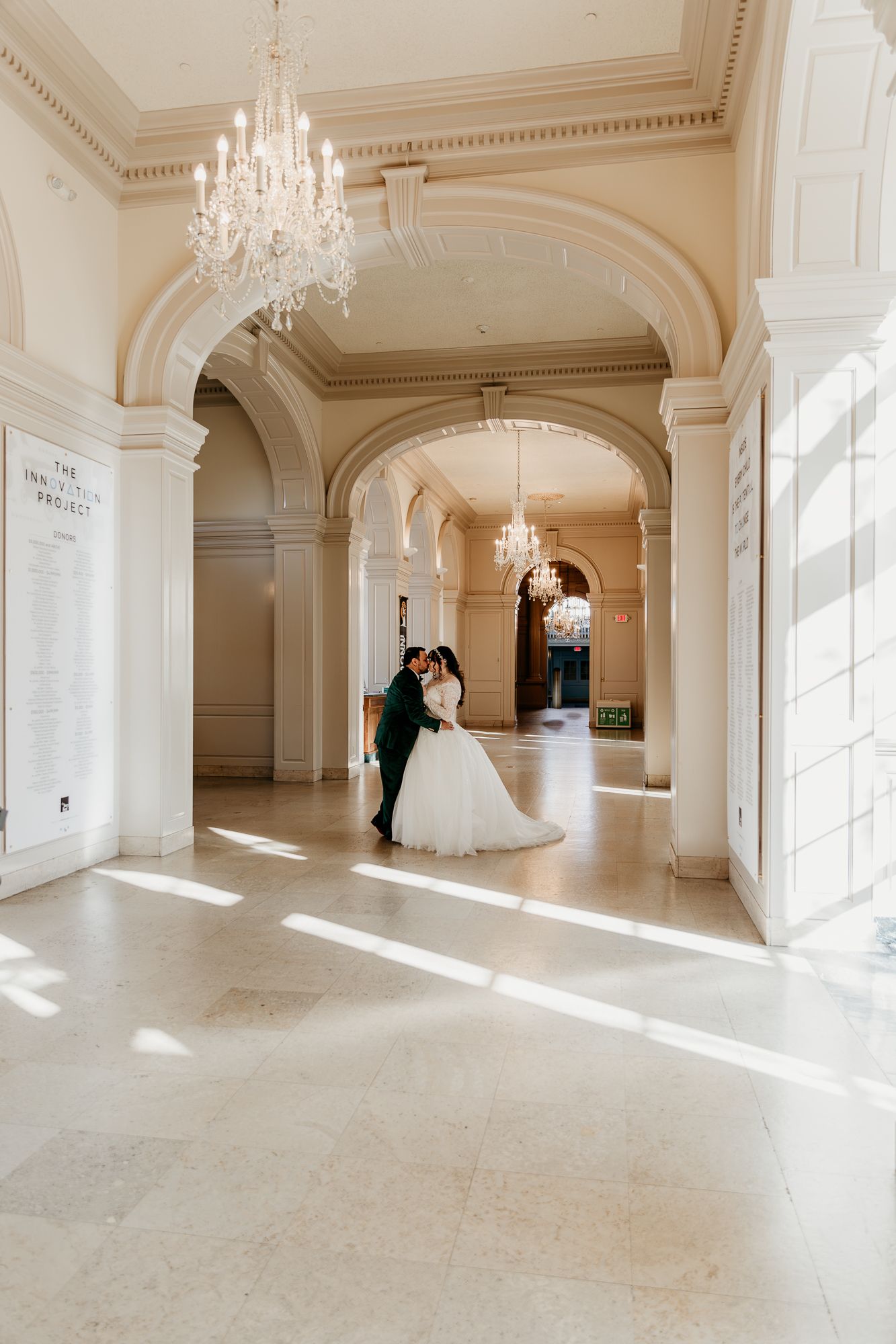 A wedding couple posing in an elegant hallway with arched columns and crystal chandeliers casting natural light.