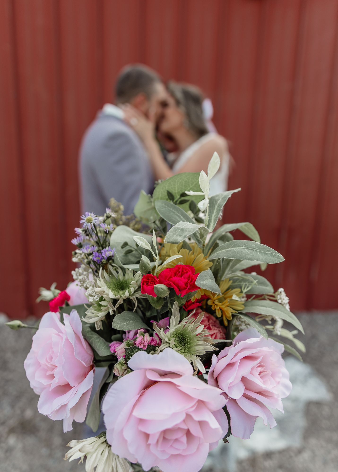 Pink and red floral bouquet with greenery in foreground of wedding couple.