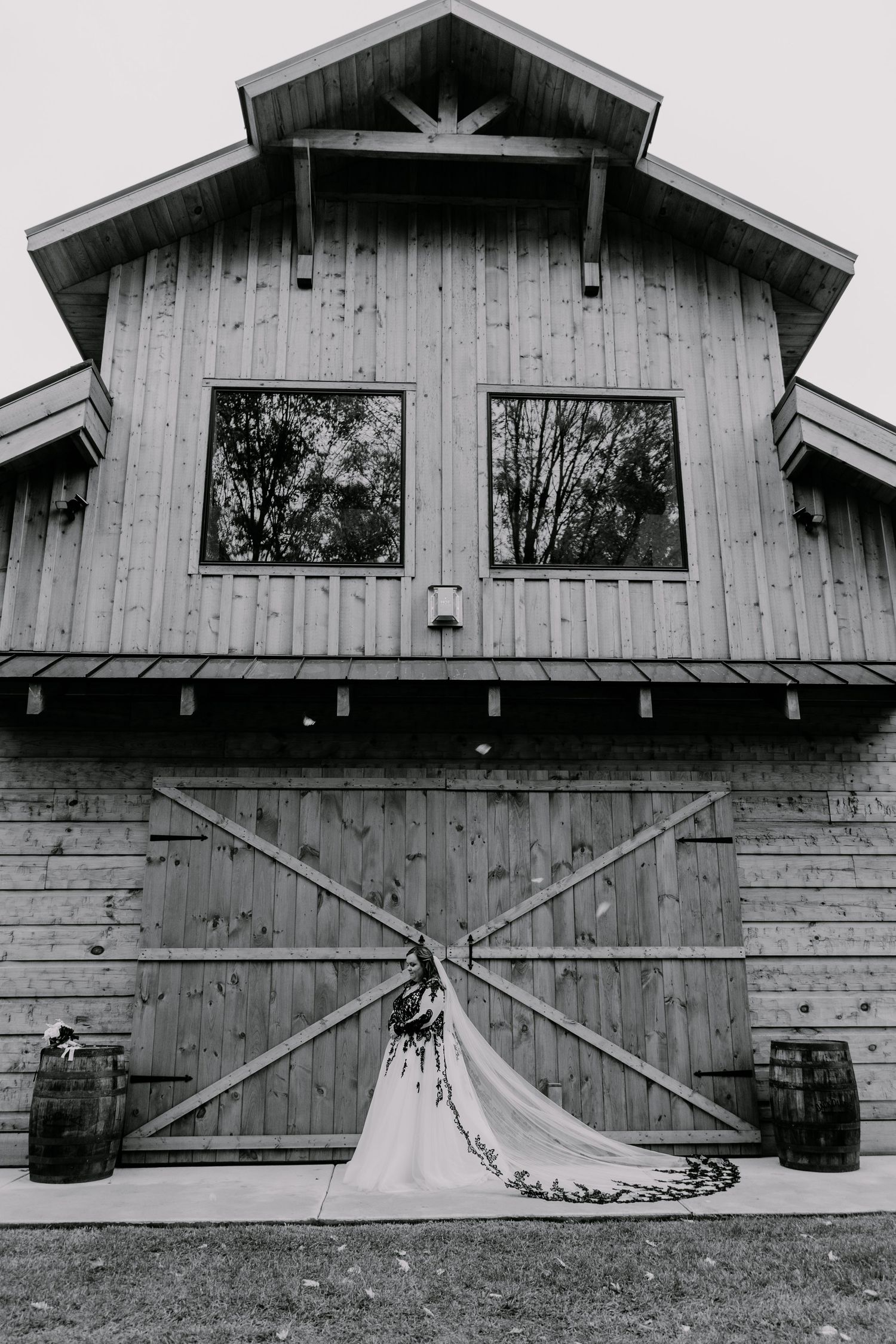 Black and white photo of a wedding dress displayed against a rustic barn door with wooden barrels.