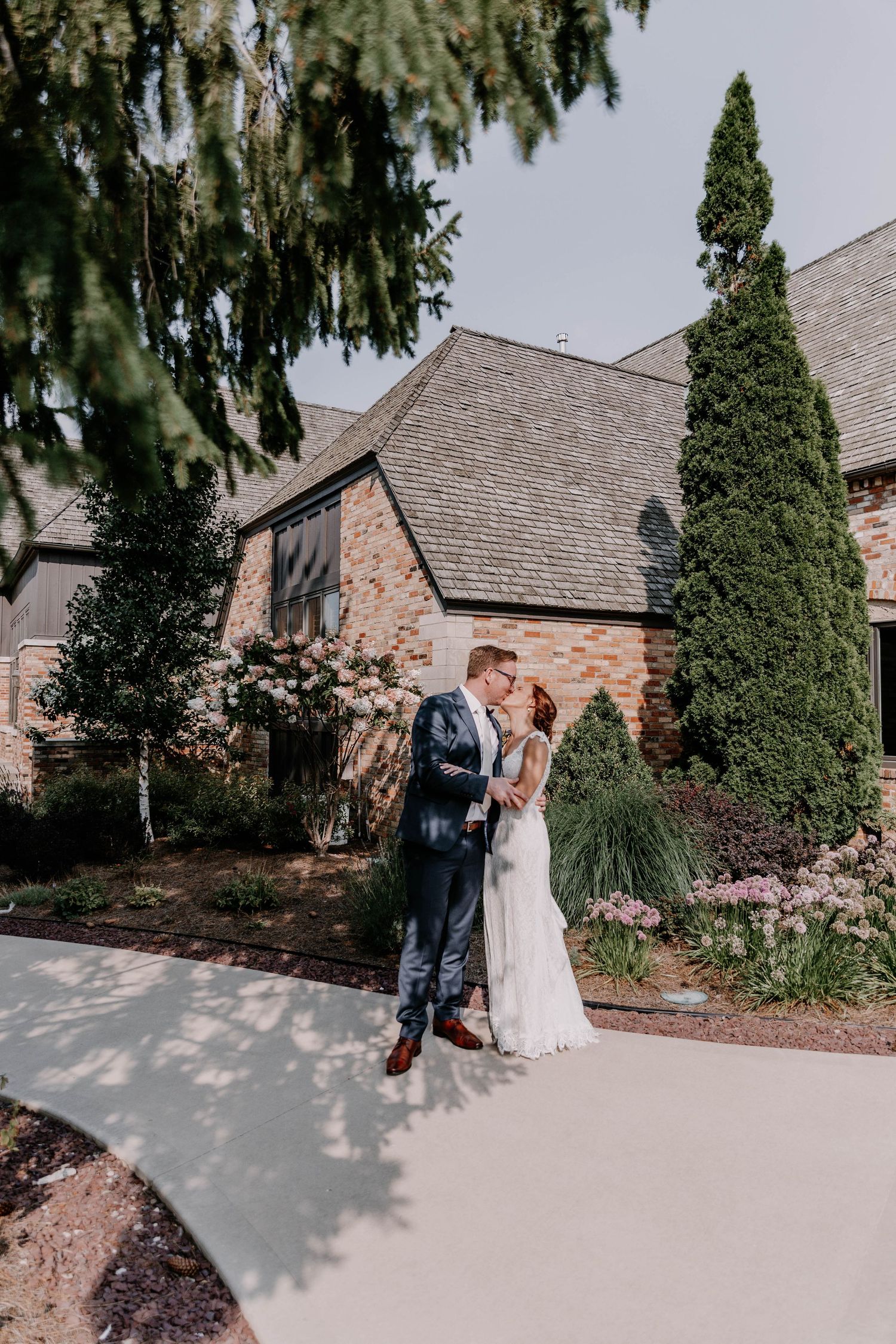 A couple shares an intimate moment outside a brick church building with tall cypress trees.