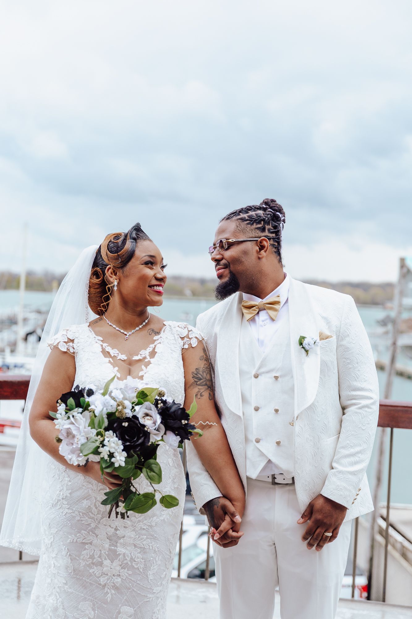 Newlyweds in elegant white attire share intimate moment on waterfront deck.