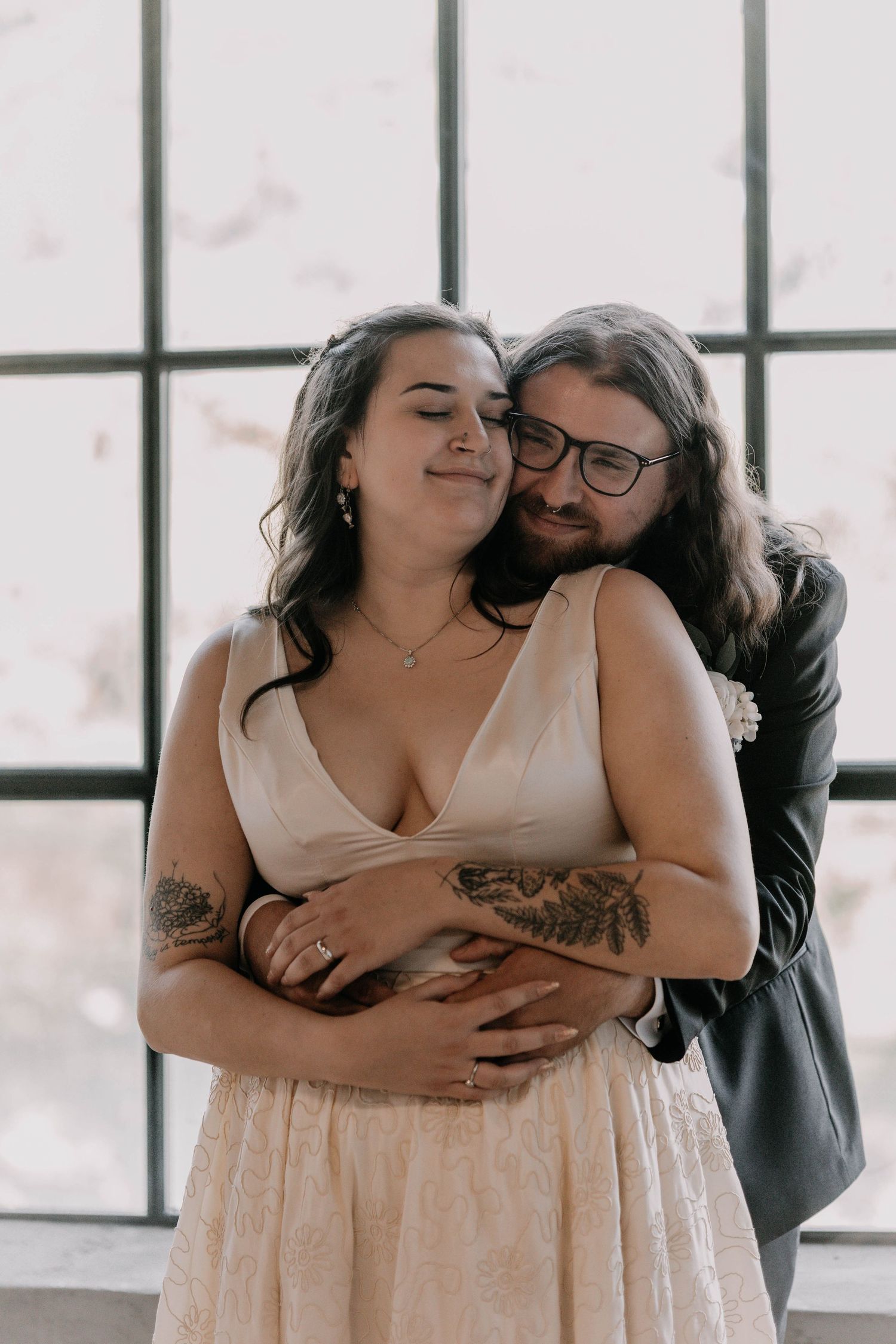 A romantic embrace between two people in formal wedding attire by a bright window.