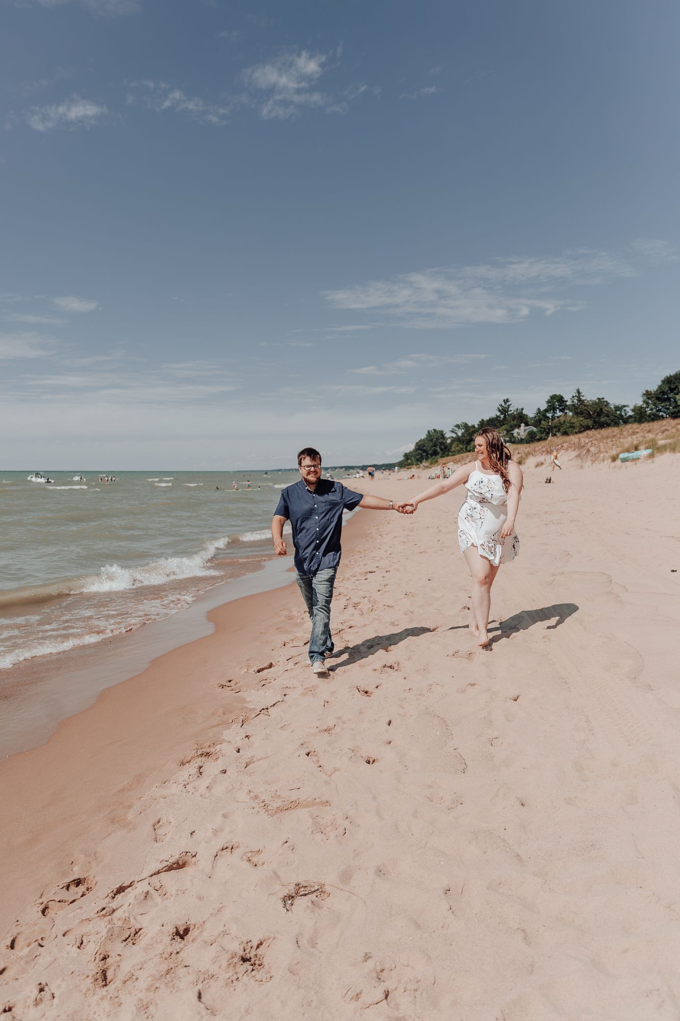 Couple holding hands while walking on sandy beach with blue waters and dunes in background.