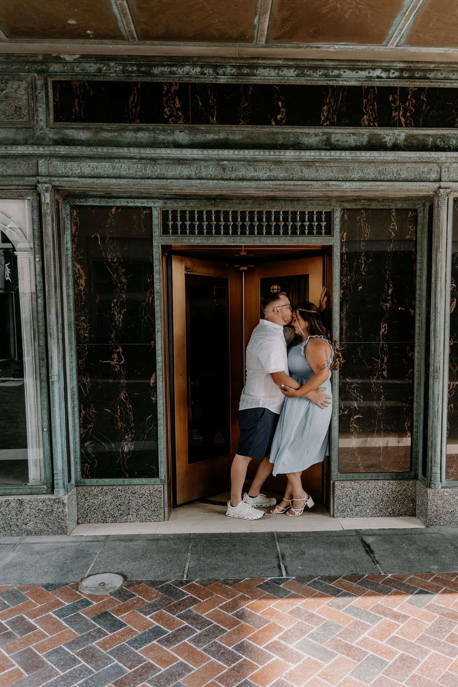 Couple embracing in vintage elevator doorway with marble walls and herringbone brick floor pattern.