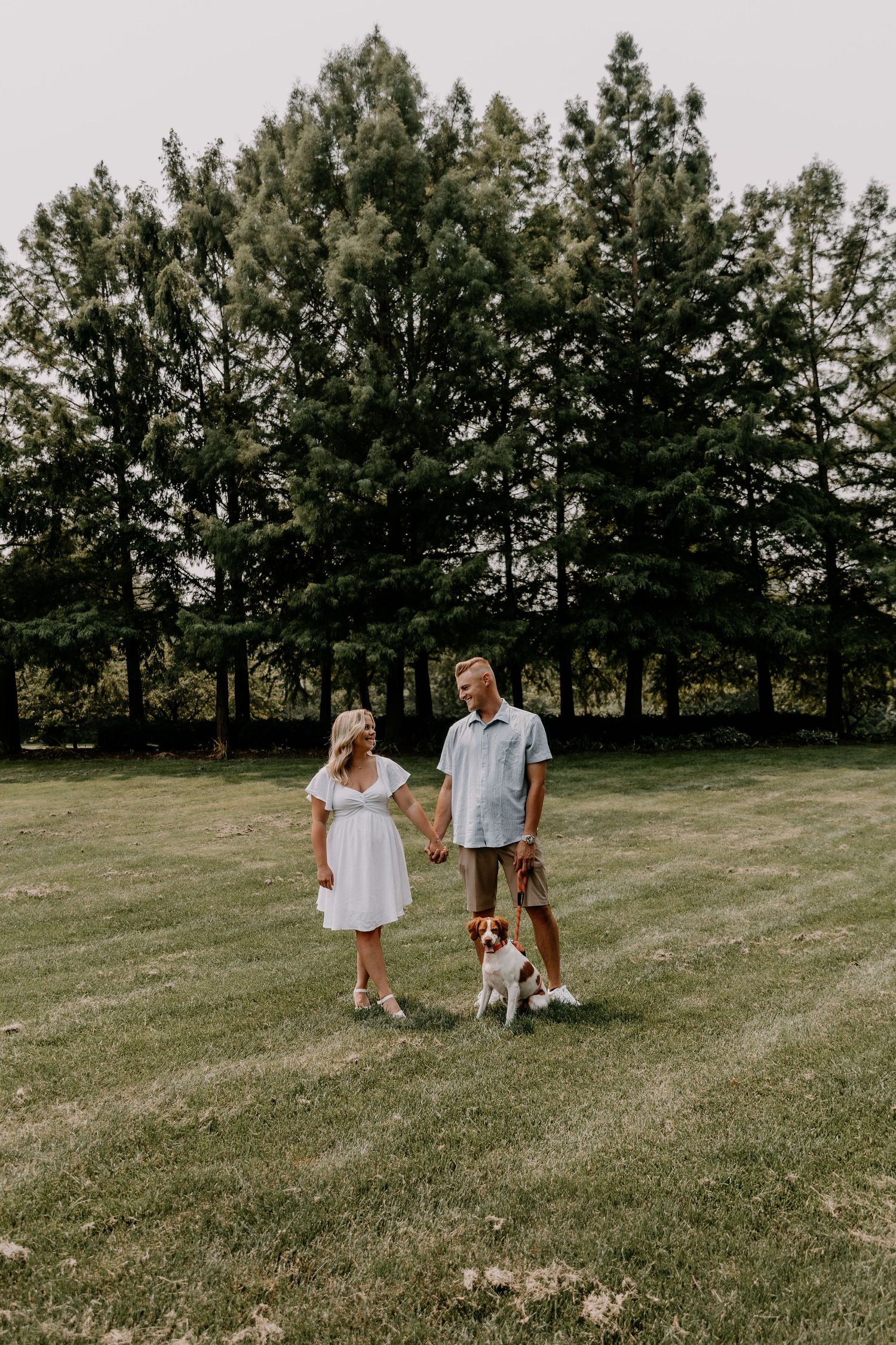 A couple and their dog share a tender moment on a grassy field with tall evergreen trees in the background.