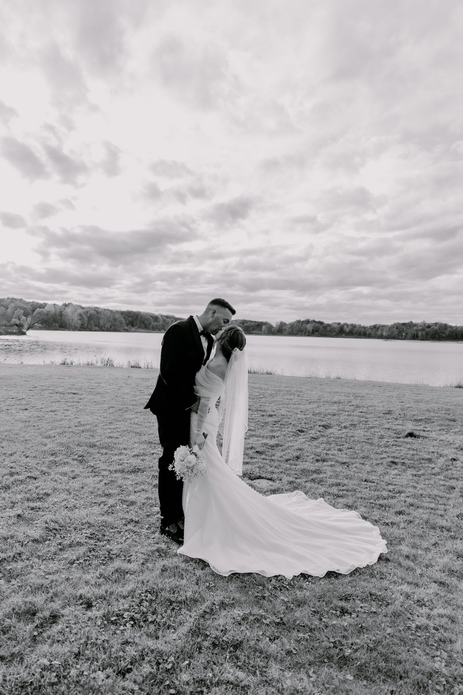 A couple in wedding attire share an intimate moment in a black and white photo on a grassy field under cloudy skies.