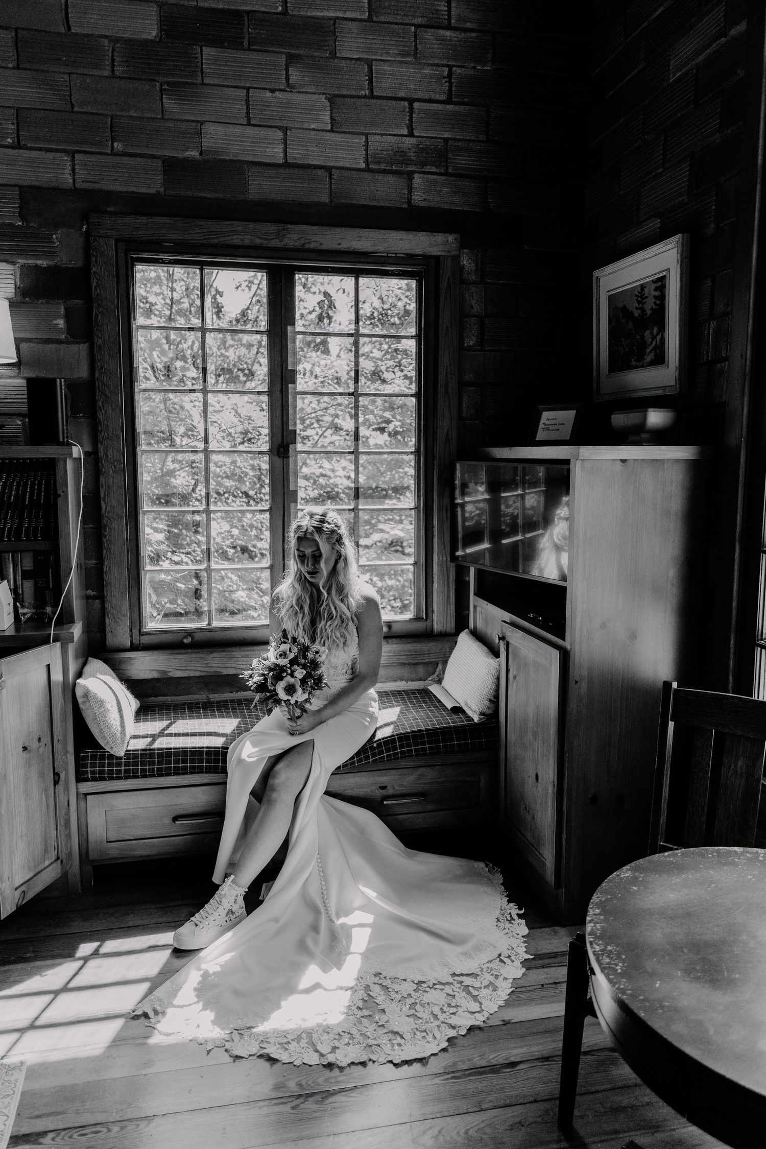 Elegant wedding scene in black and white showing a flowing dress by a window in a rustic brick interior.