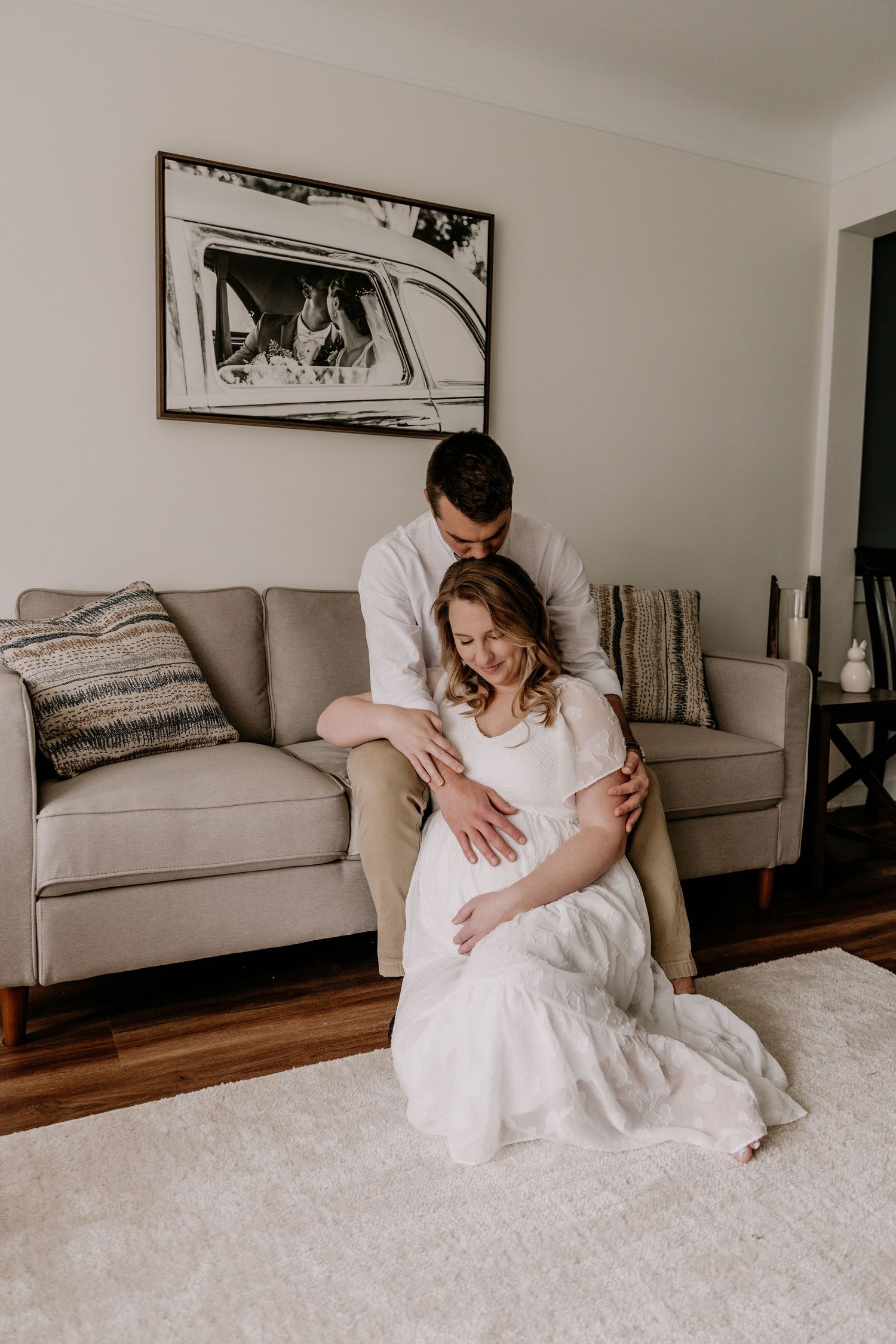 A man tenderly embraces a pregnant woman in a white dress as they sit together on a cozy living room floor.