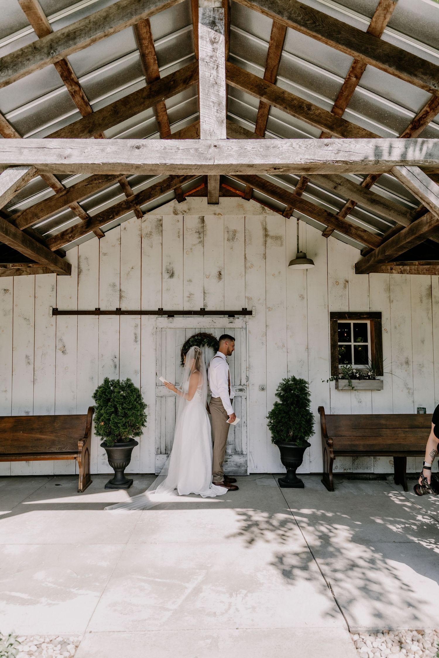 Bride and groom stand together at rustic white barn wedding venue with wooden beams and potted topiaries.