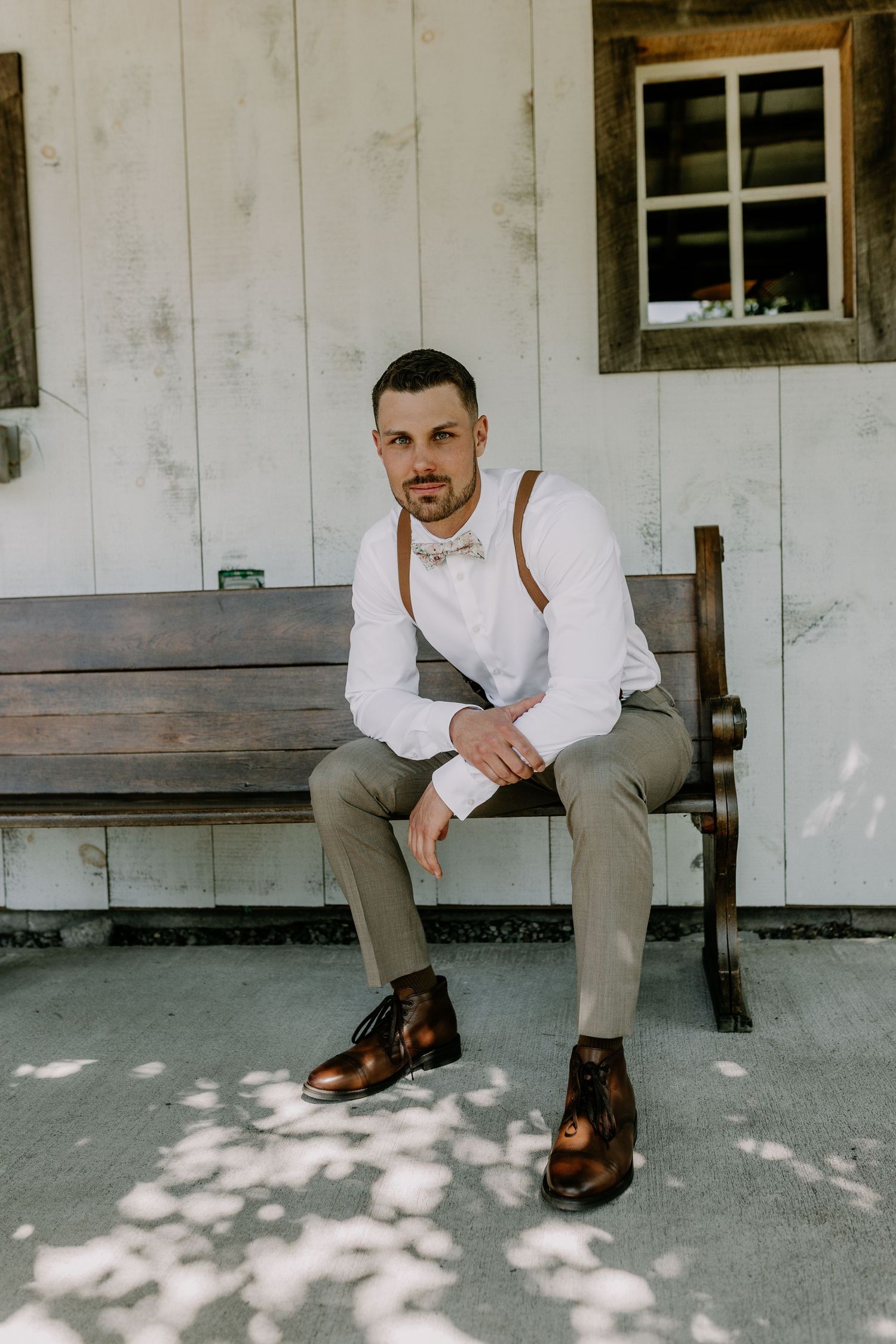 A well-dressed groom sits on a wooden bench wearing suspenders, bow tie, white shirt, and brown leather boots.