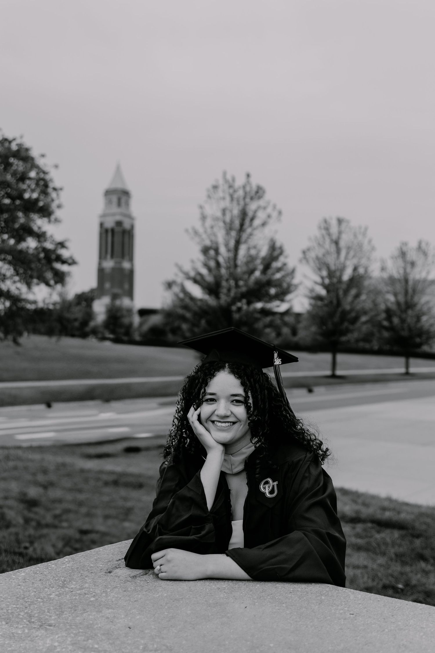 Black and white graduation photo showing graduate in cap and gown sitting outdoors with church bell tower in background.