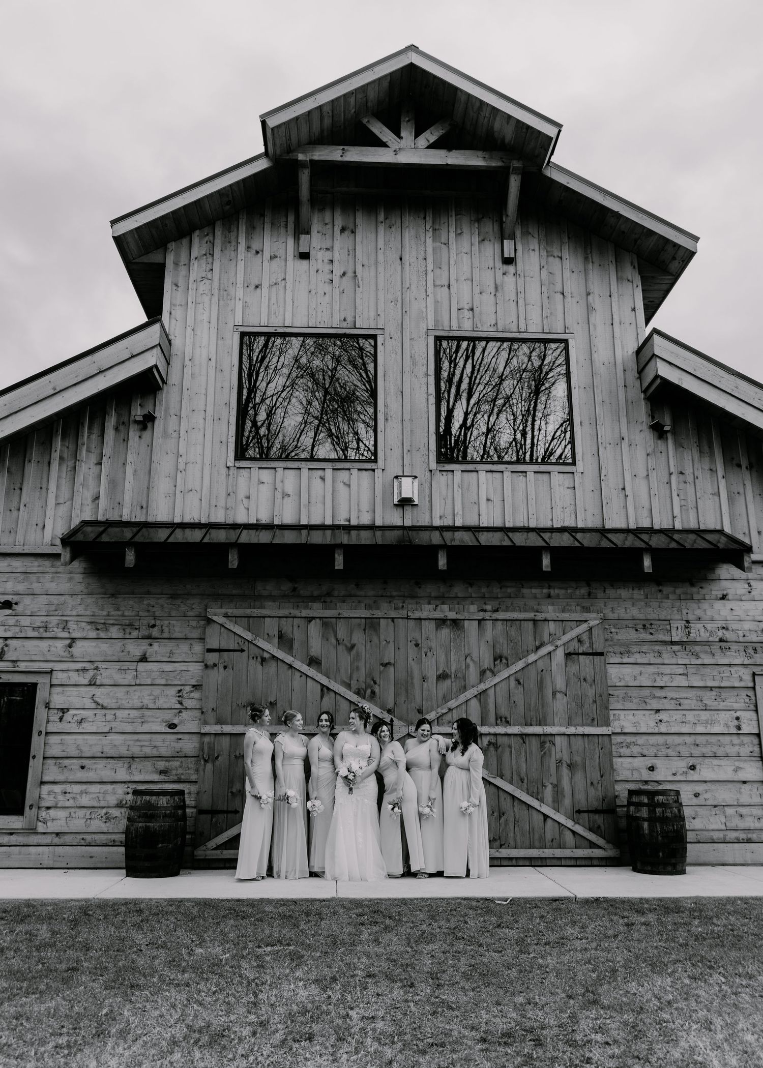 Bridal party in light colored dresses poses in front of rustic barn venue.