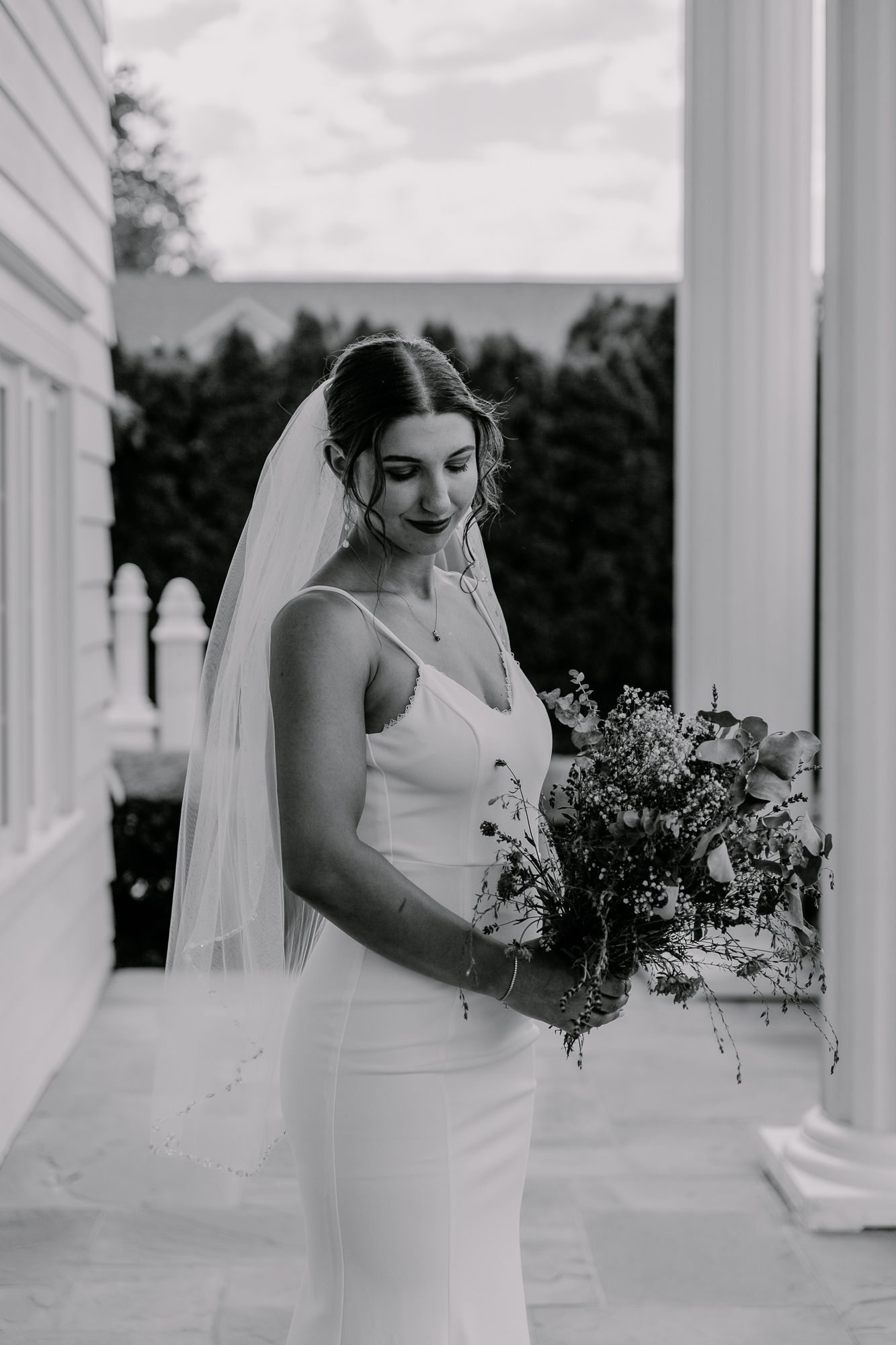 A bride in a fitted white dress holds a wildflower bouquet while standing by white columns.